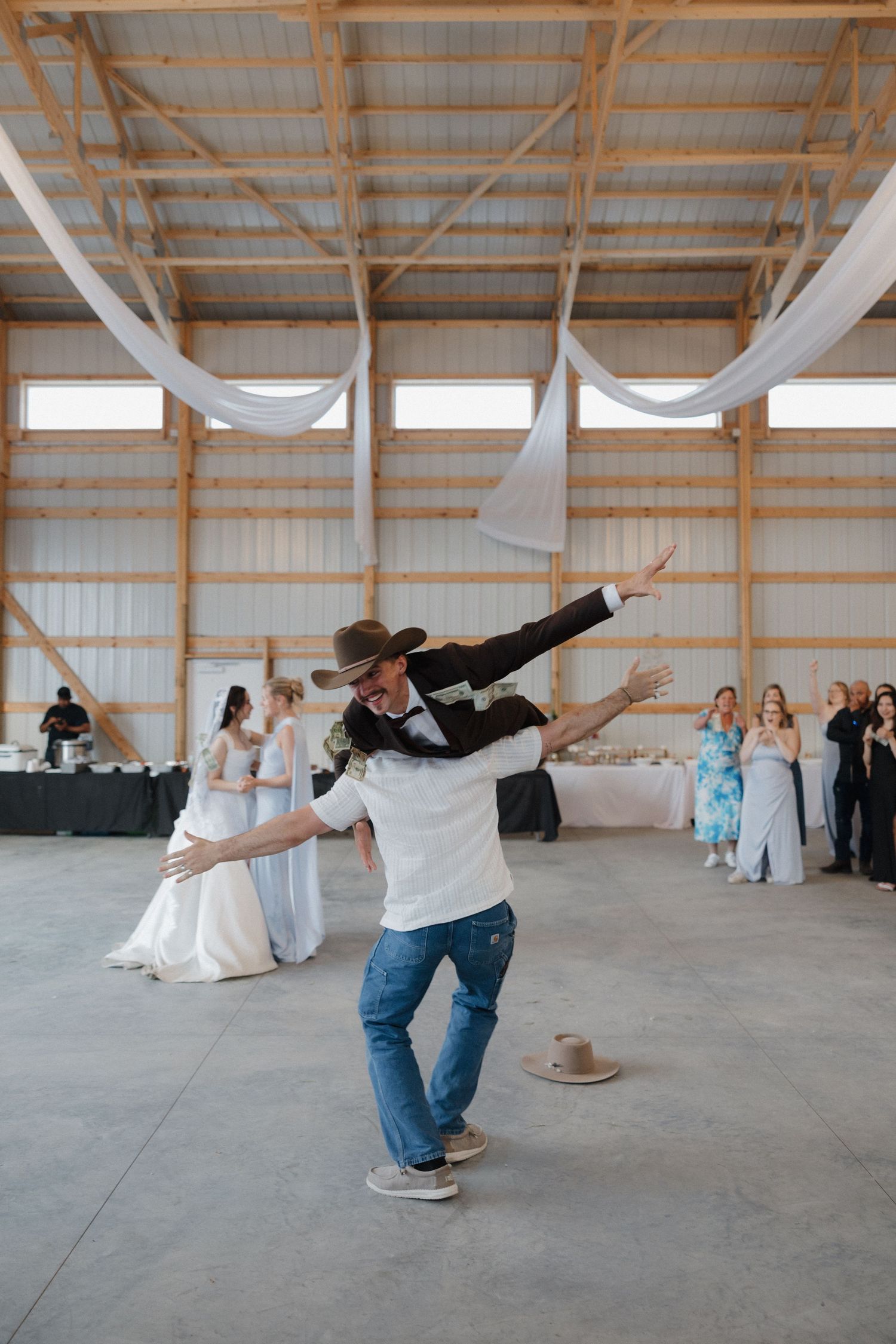 A couple performs a playful dance dip sequence in a rustic barn venue with white drapes and wooden beams overhead.
