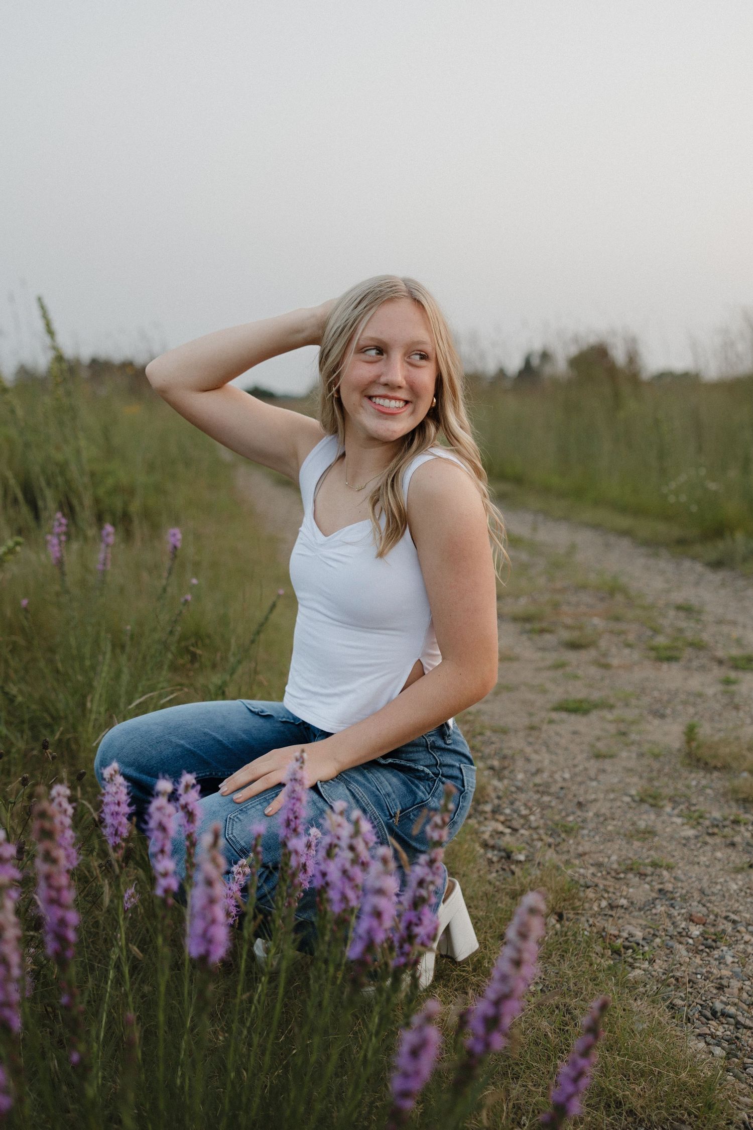 Smiling blonde teenage girl posing among purple wildflowers on a gravel path in a green field.