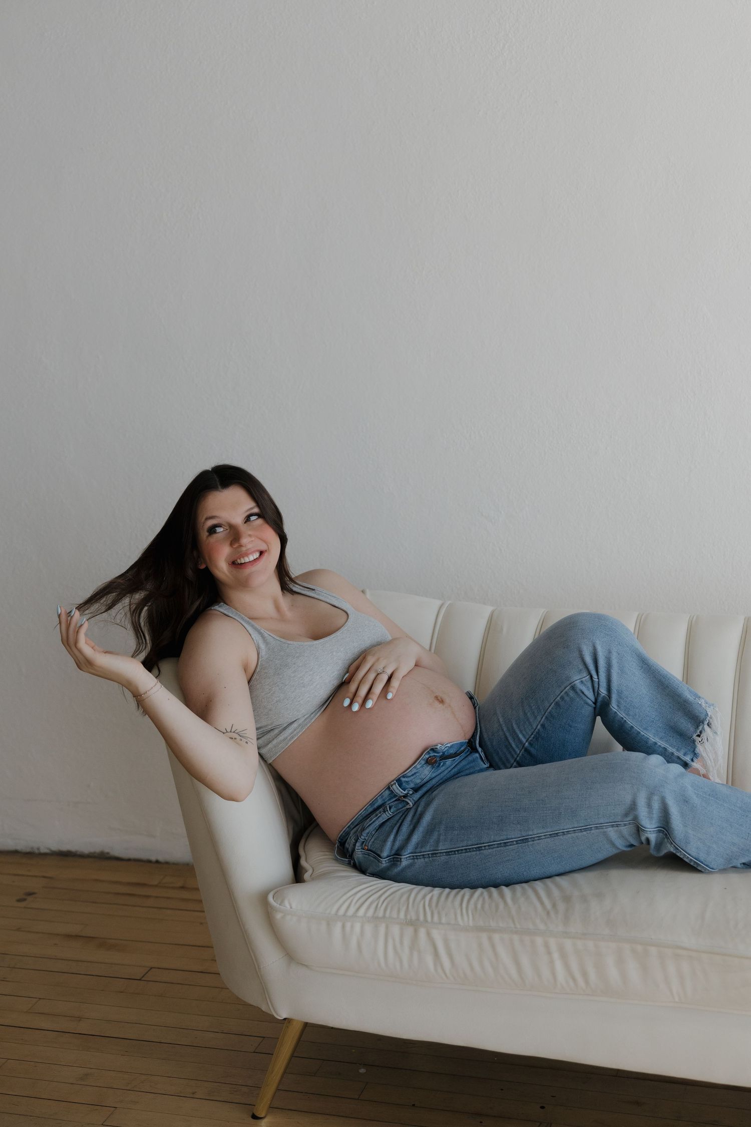 Smiling pregnant woman relaxing on cream sofa in gray tank top and jeans, gently holding her baby bump.