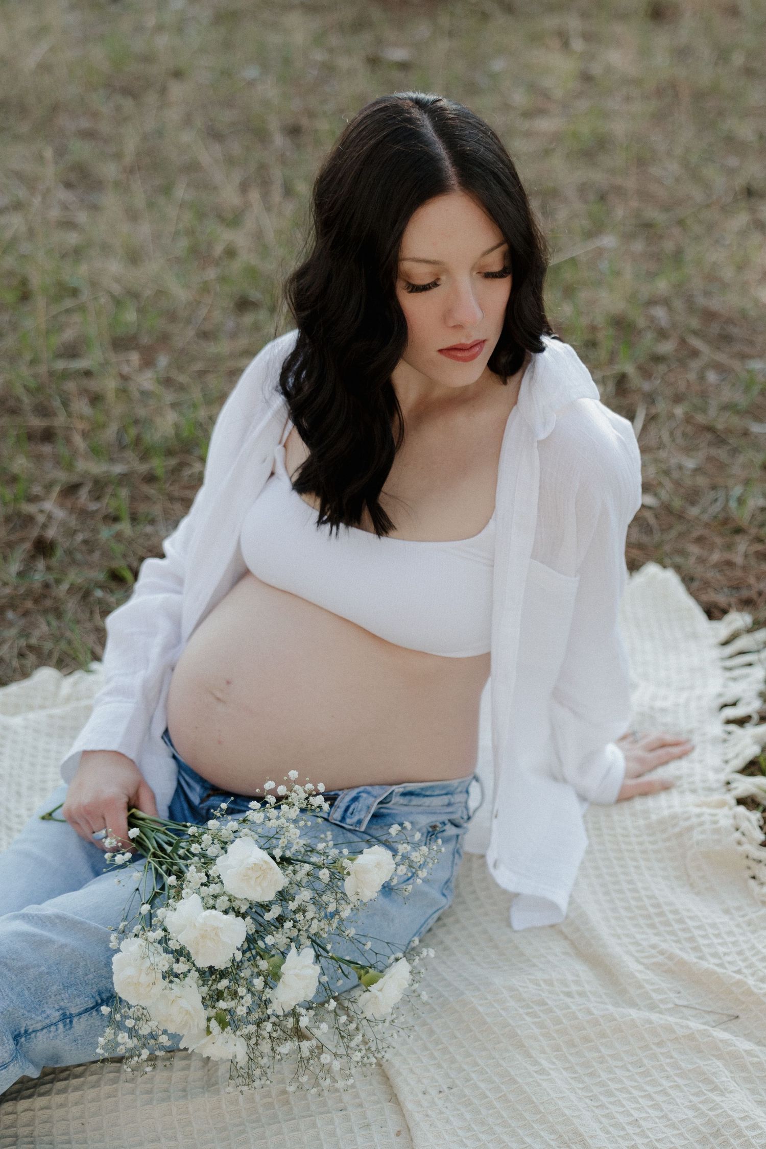 Pregnant woman in white shirt and jeans sitting on blanket, holding white flower bouquet near baby bump.