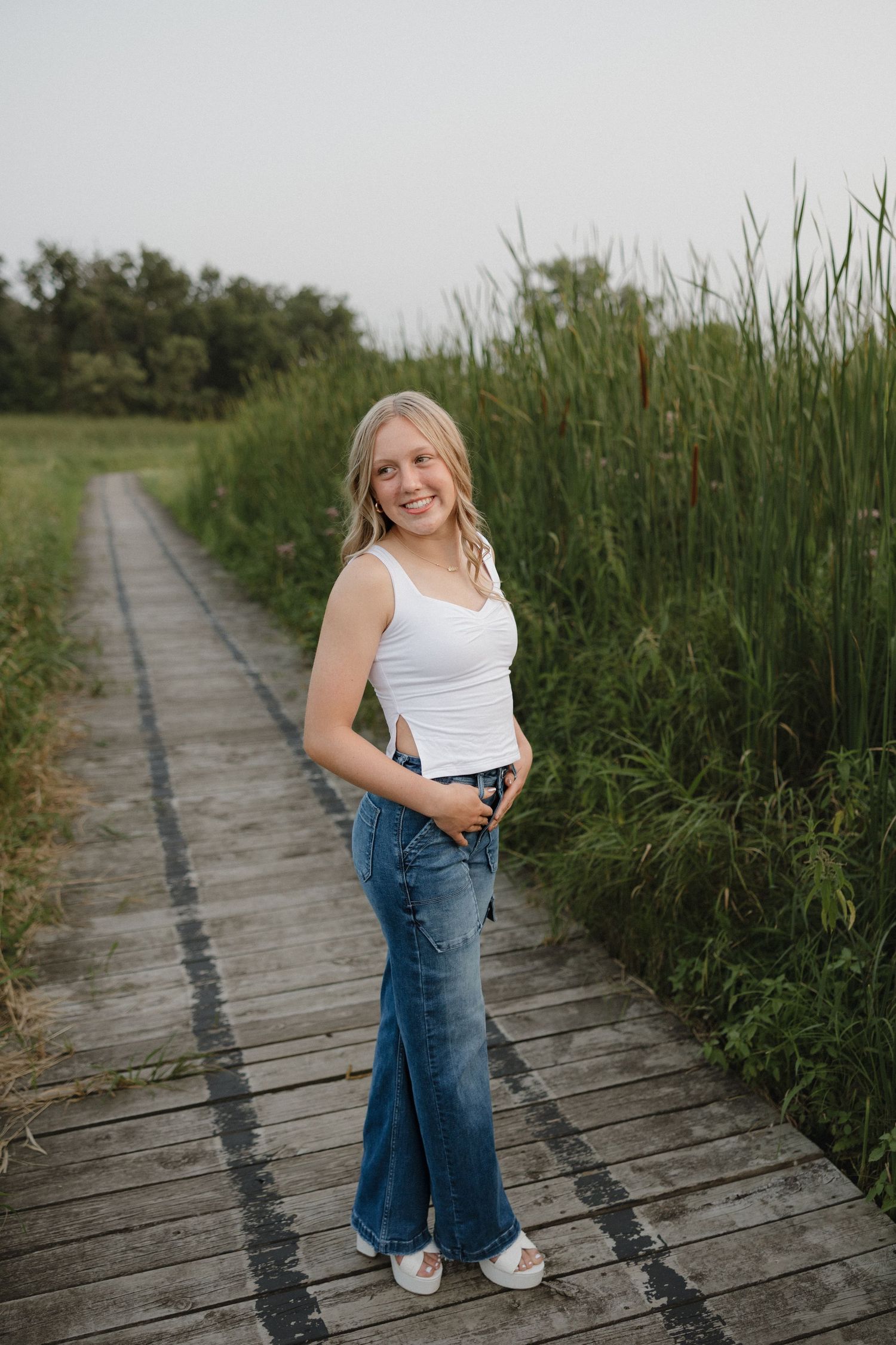 Smiling blonde woman in white tank top and wide-leg jeans standing on wooden boardwalk surrounded by tall reeds.