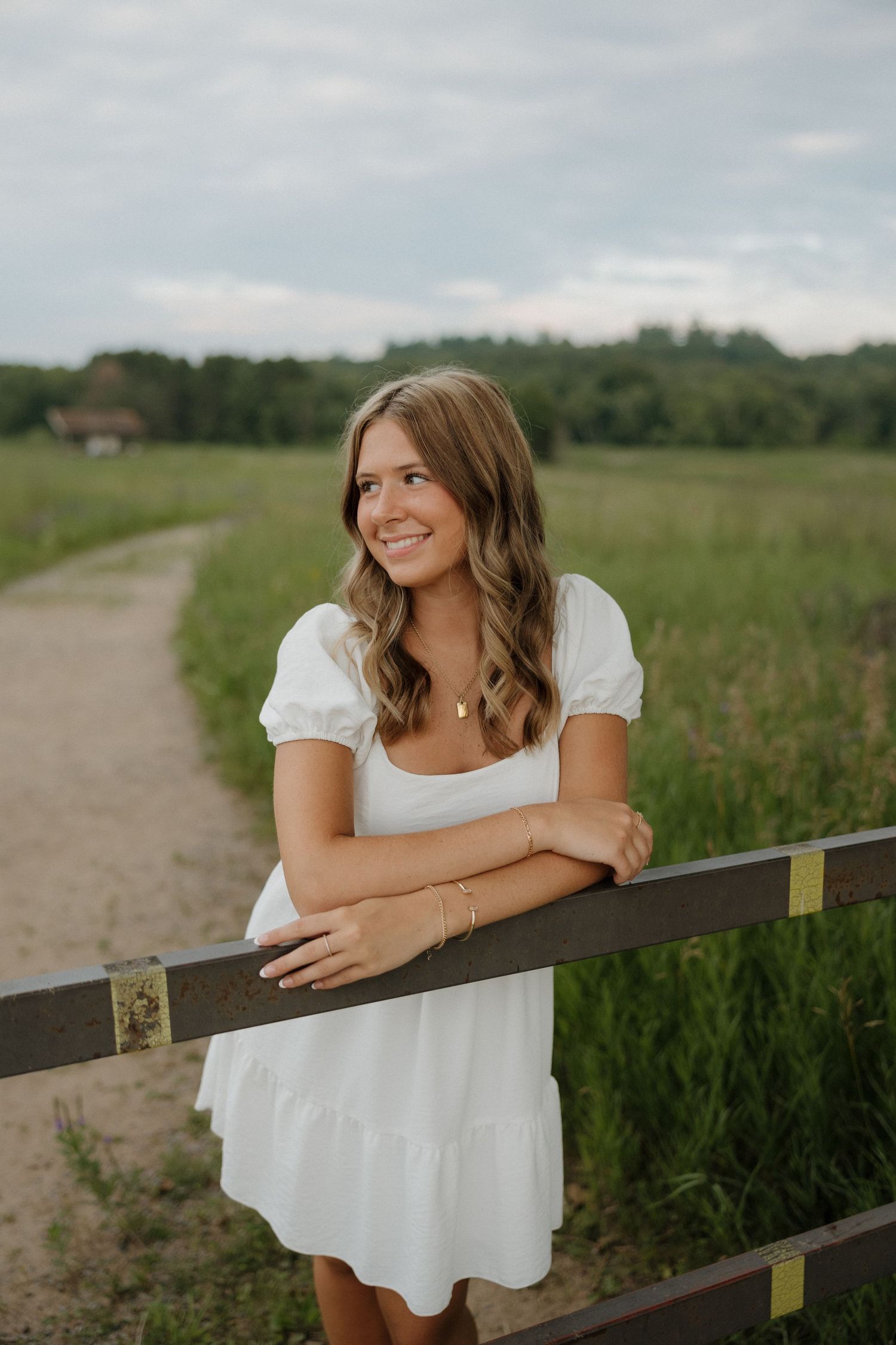 Smiling young woman in white dress leaning on a wooden fence rail along a scenic field path.
