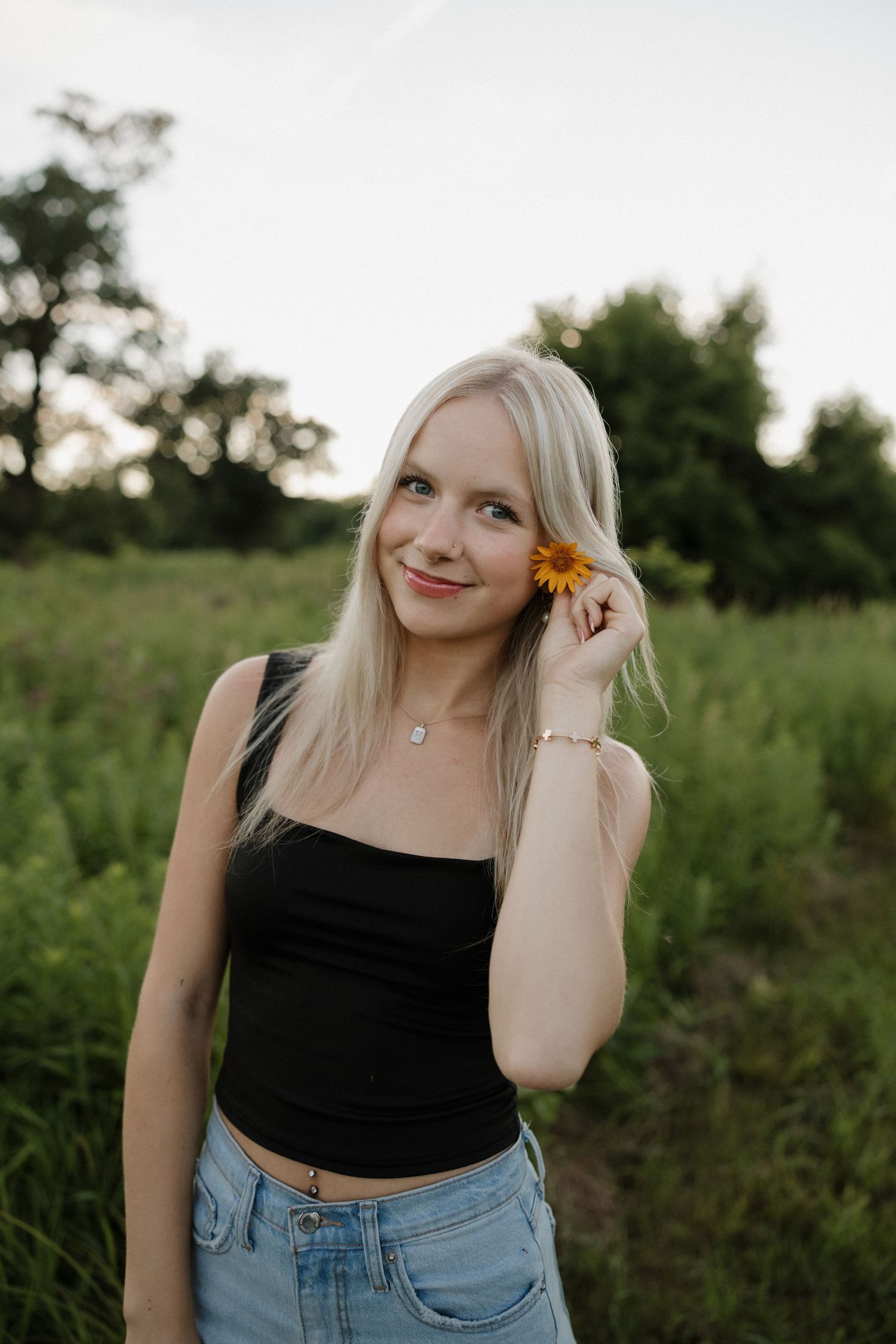Blonde woman in black crop top holding a yellow sunflower near her face in a green field.