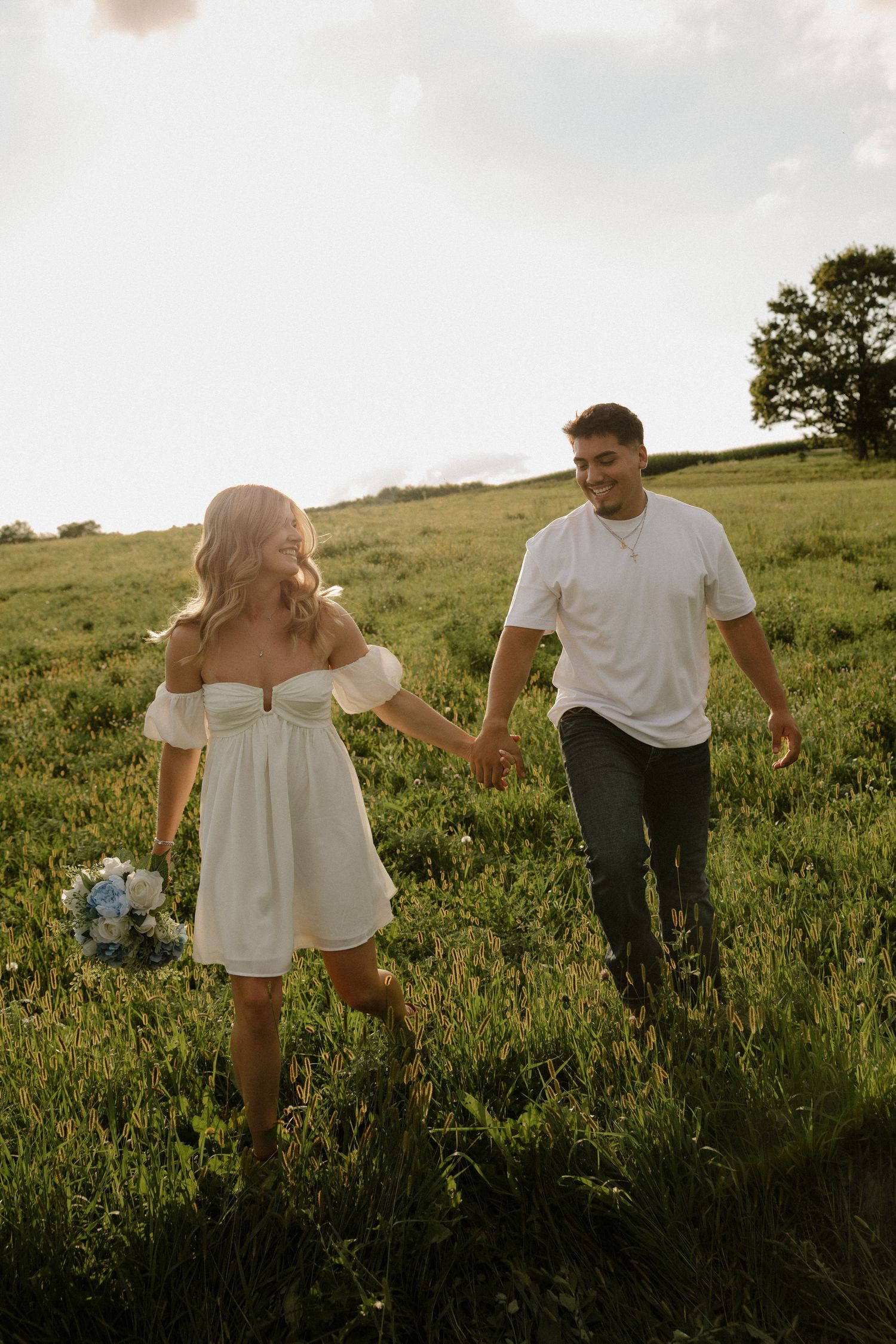 Happy couple holding hands and walking through a lush green meadow at golden hour, woman holding blue flowers.