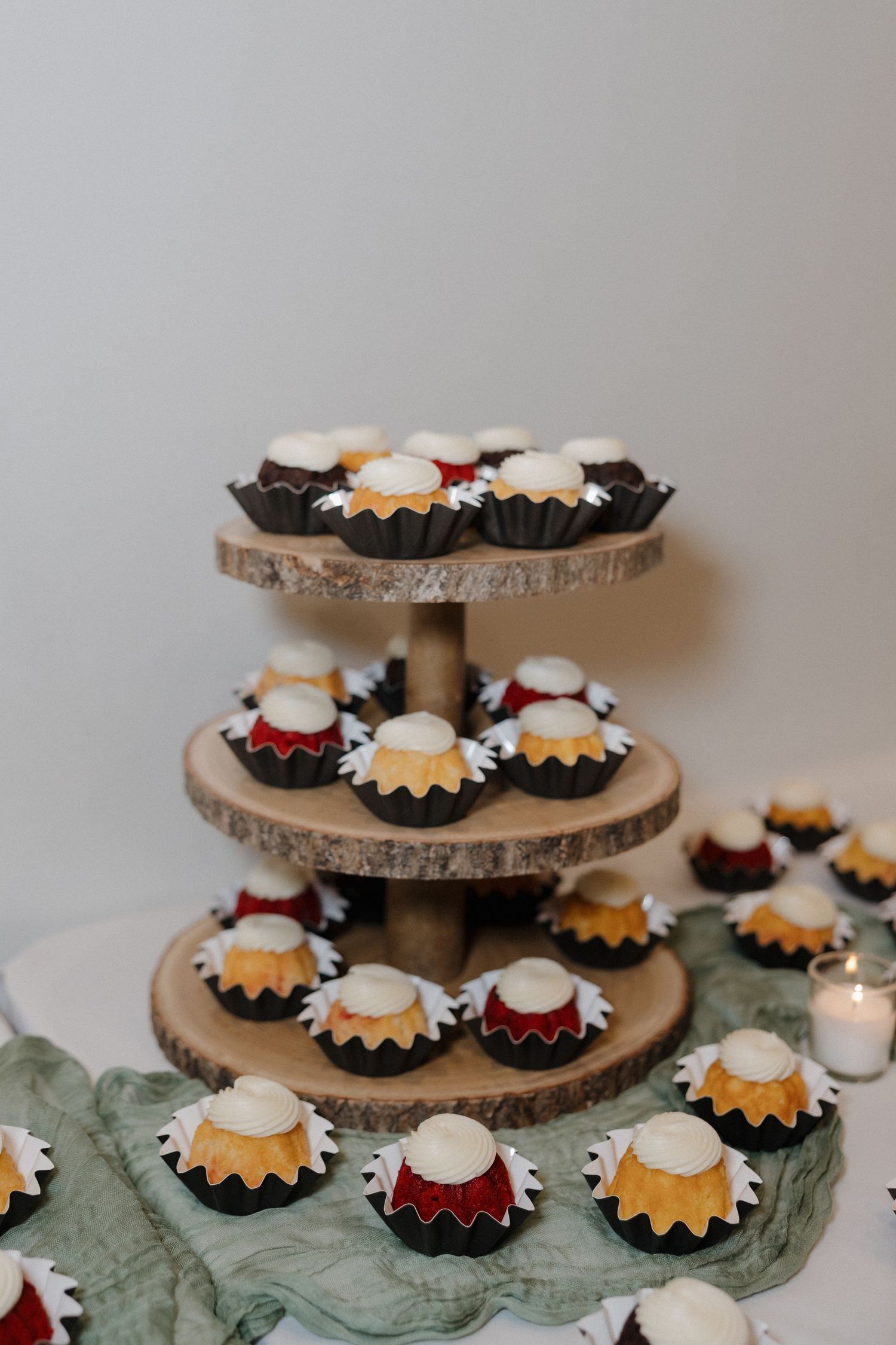 Multi-tiered rustic wood cake stand displaying mini bundt cakes with white frosting on sage green fabric.