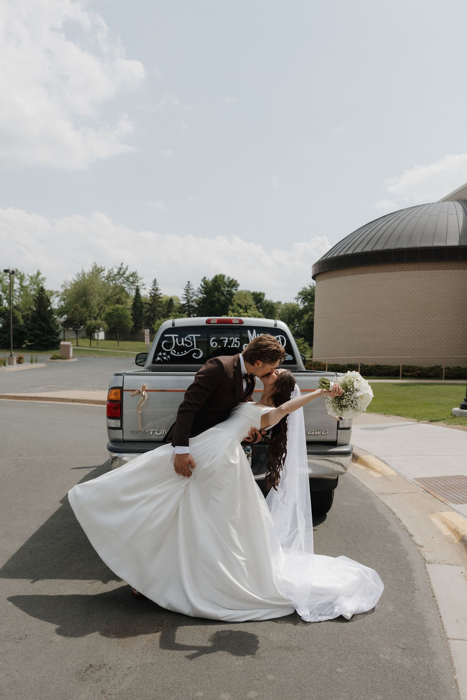 A wedding couple performs a dramatic dip pose in a parking lot, with the bride's white dress flowing.