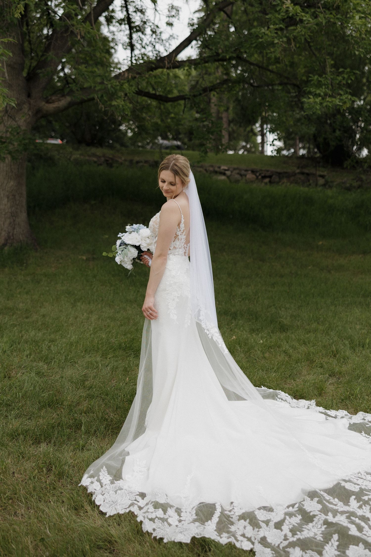 Bride in lace wedding gown with long cathedral veil holding white bouquet, posing in a garden.