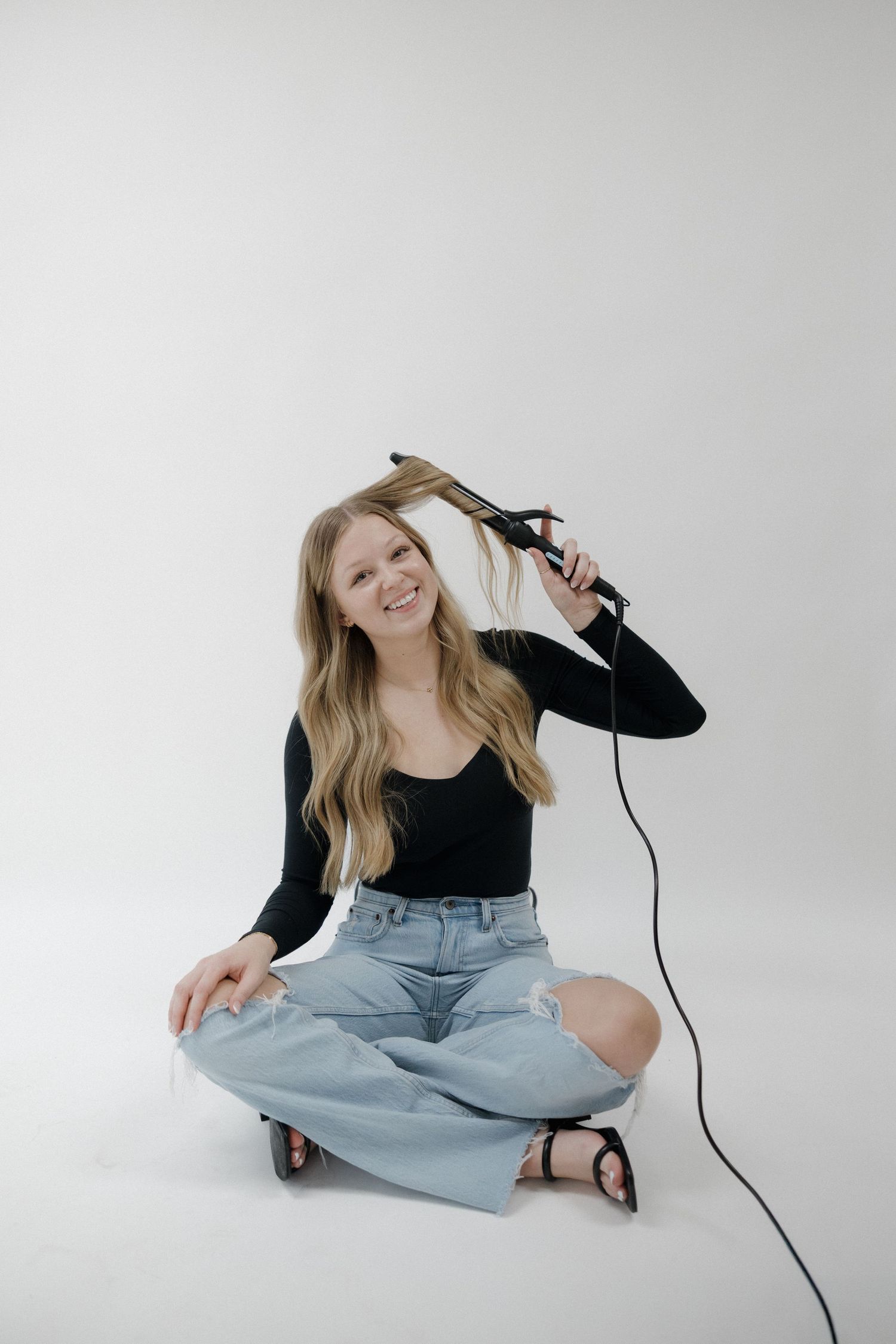 Smiling blonde woman sitting cross-legged using a flat iron hair straightener on white background.