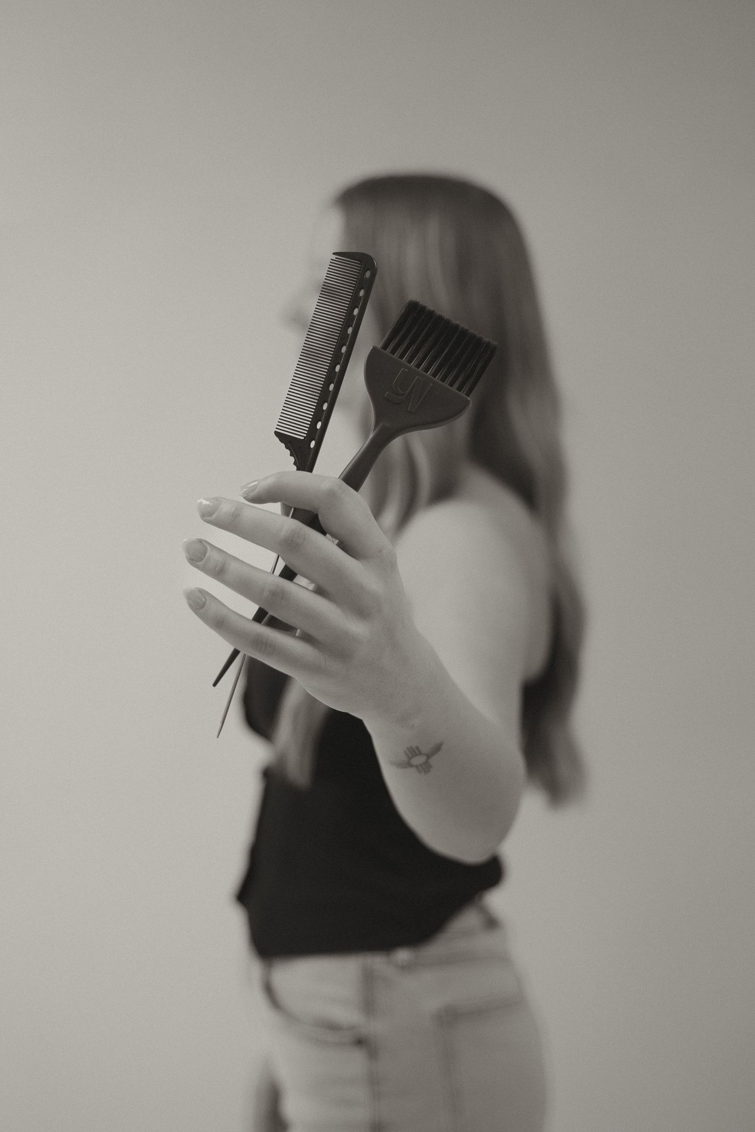 Black and white photo of woman holding hair combs in front of her face with tattoo visible on wrist.