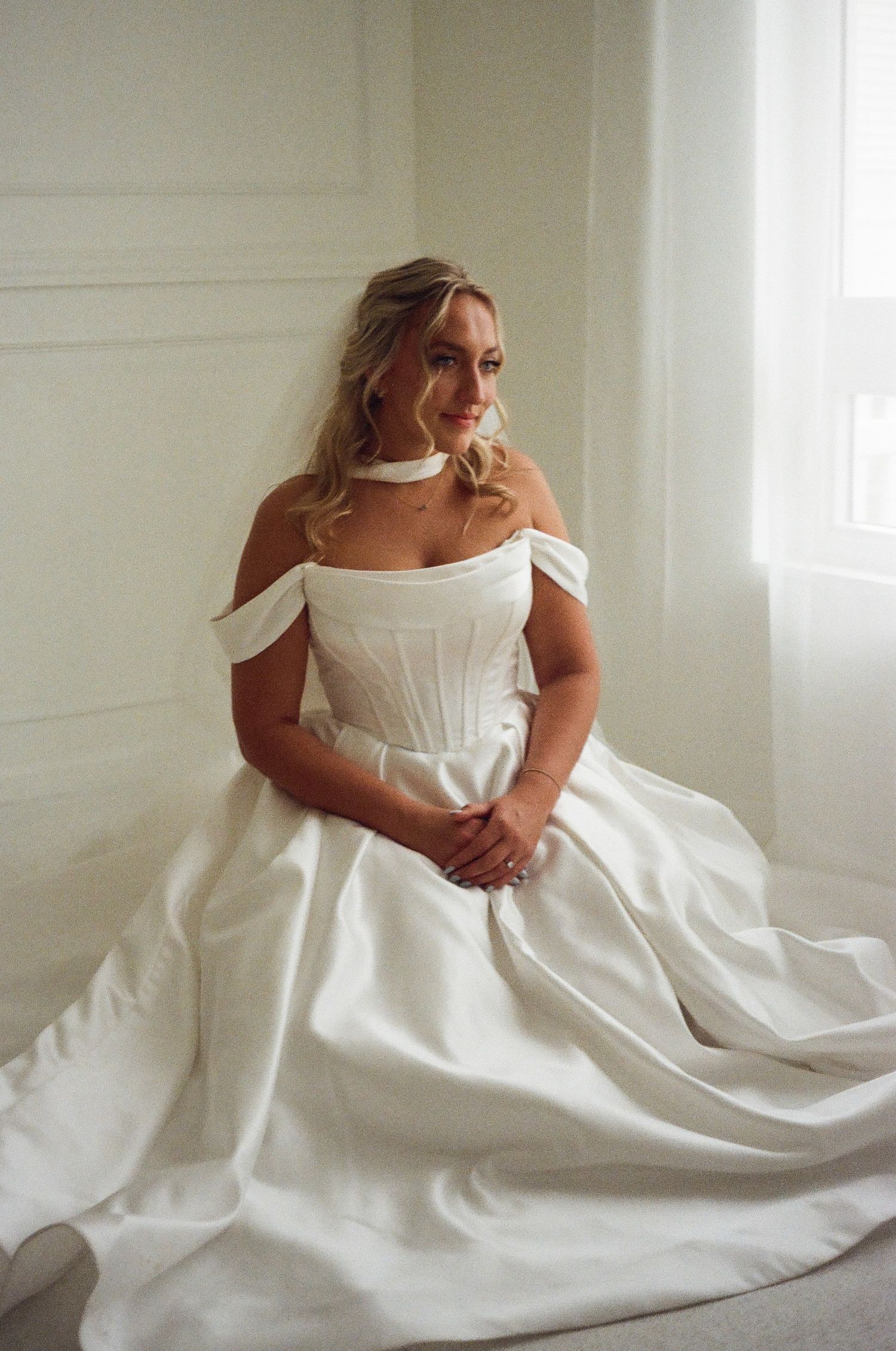 Bride in an off-shoulder white ball gown with corset bodice sits gracefully in a softly lit room.