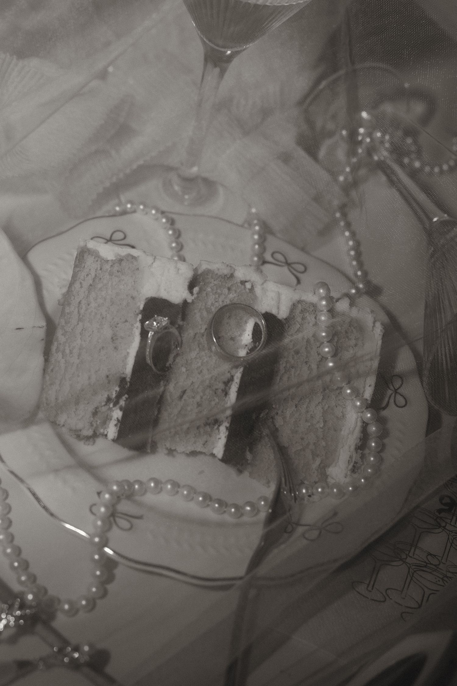 Black and white photo of wedding rings resting on cake slices with pearl necklace decoration.