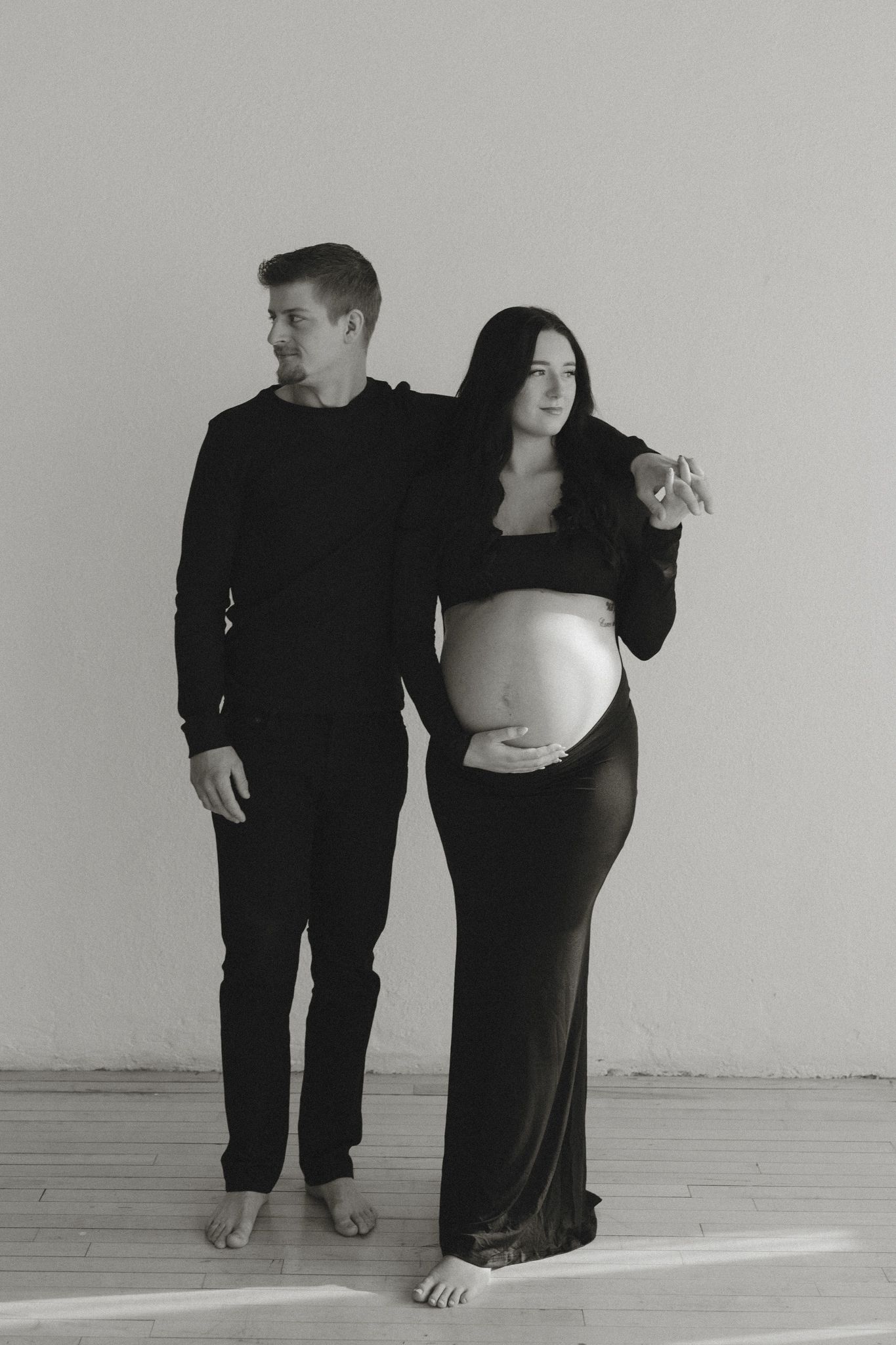 Black and white maternity photo of pregnant woman in black dress with partner in studio.