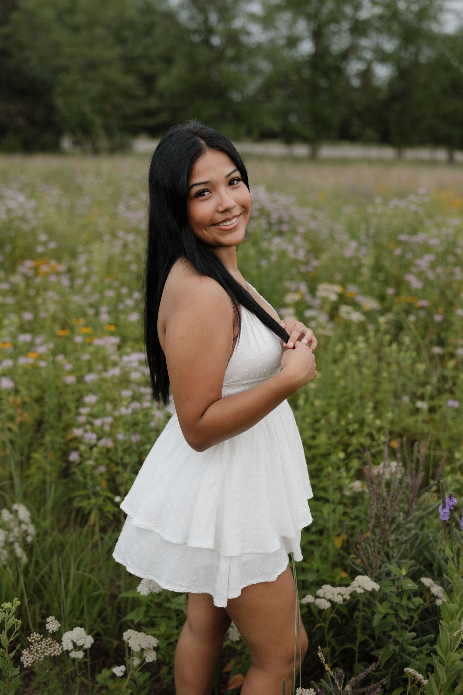 Smiling young woman in white ruffle mini dress posing in colorful wildflower field with trees behind.