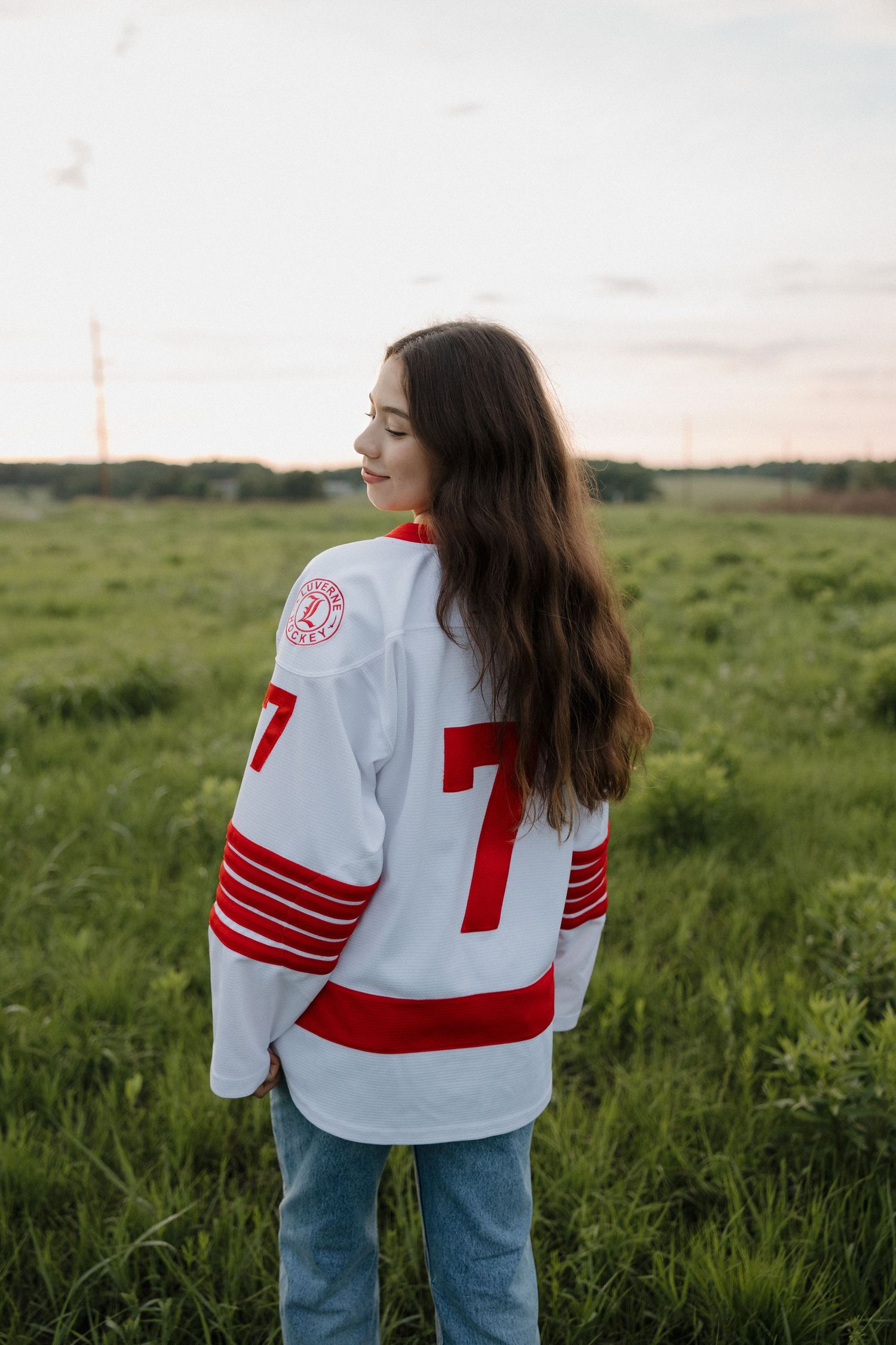 Young woman wearing a white and red hockey jersey number 7 standing in a green field at sunset.