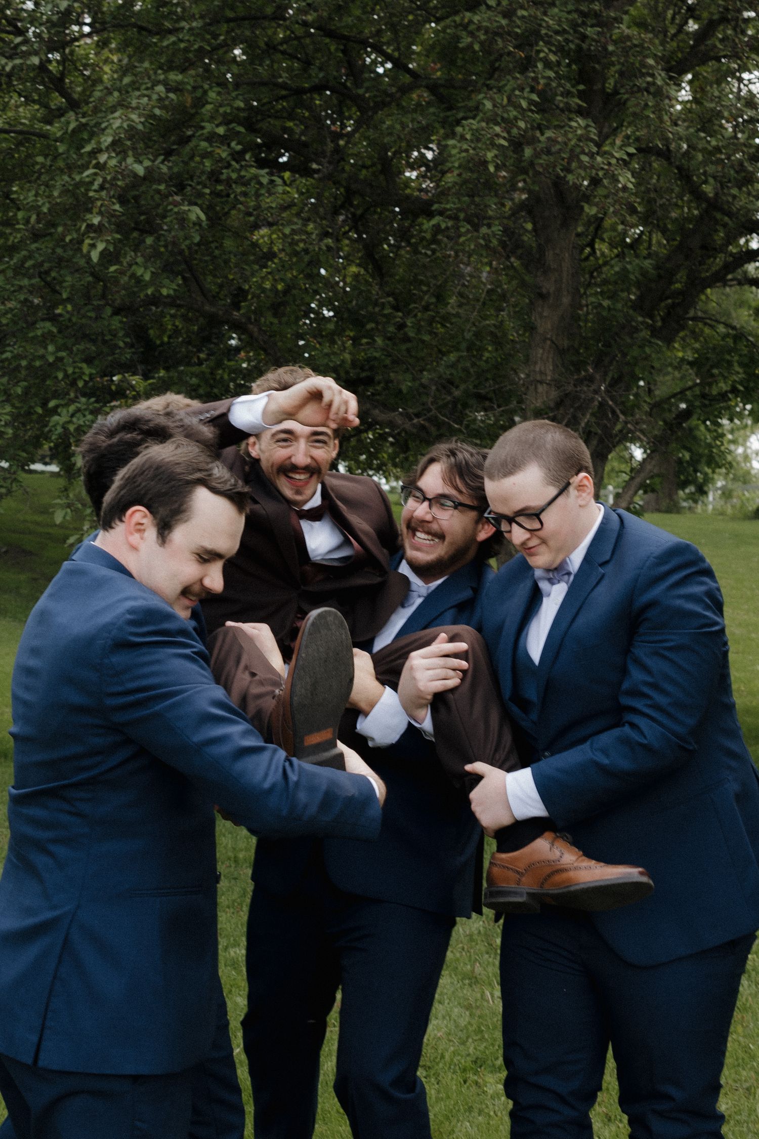 A group of groomsmen in navy suits playfully lift and carry one of their friends during a wedding photoshoot outdoors.