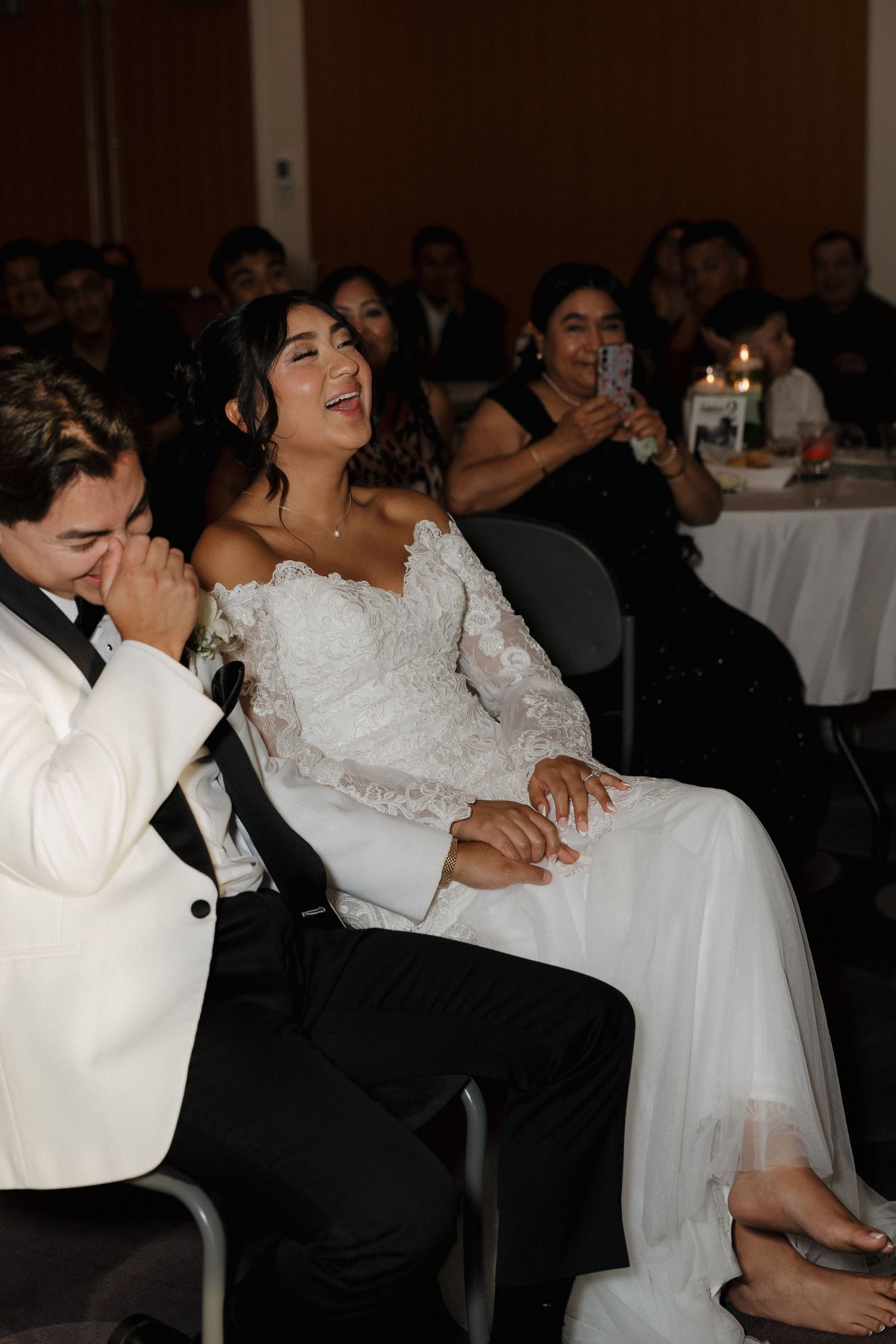 Newlyweds share a joyful moment while seated at their wedding reception with guests in the background.