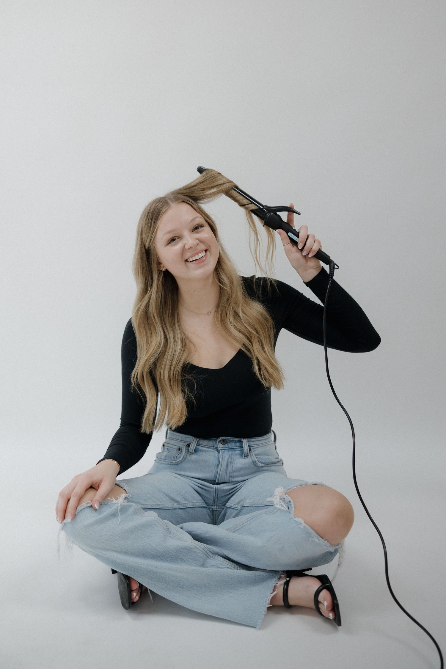 Smiling blonde woman sitting cross-legged on the floor using a hair straightener on her long hair.