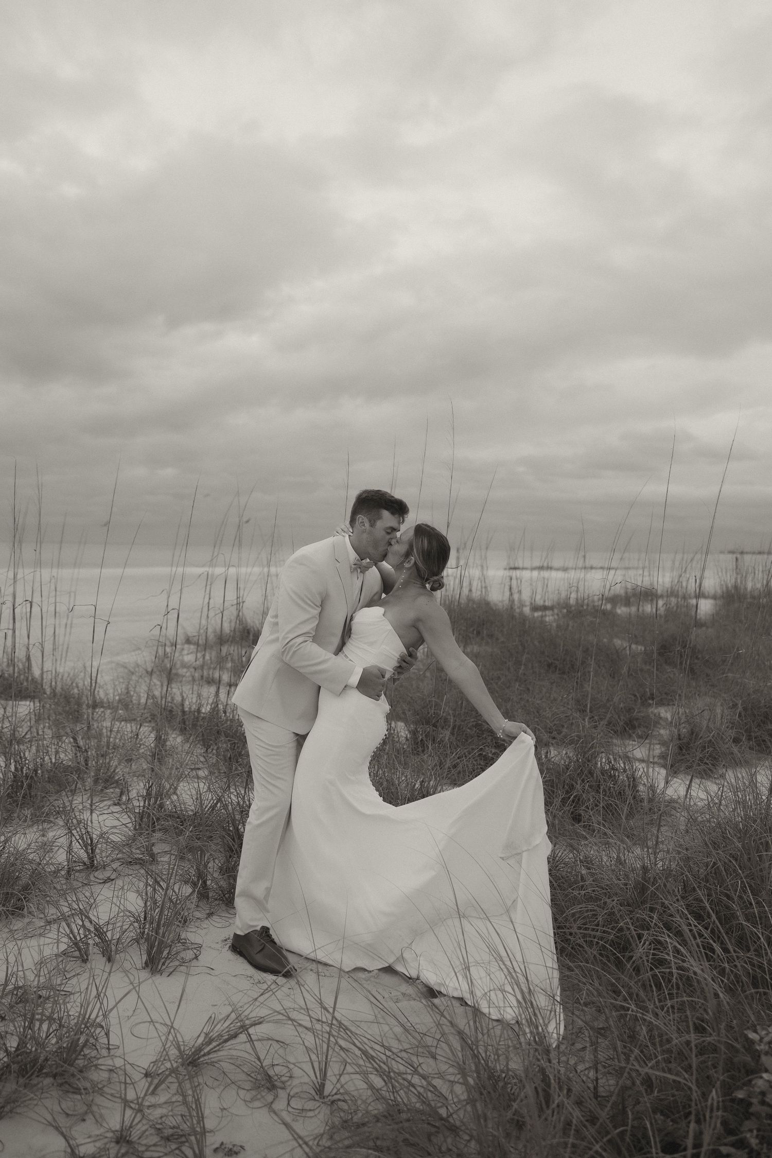 Black and white photo of a bride and groom sharing a kiss on sandy beach dunes with ocean and cloudy sky backdrop.