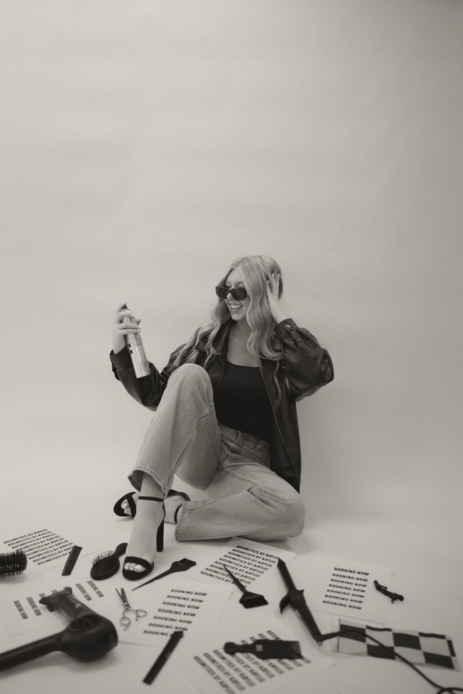 Black and white photo of blonde woman in leather jacket sitting among hair tools and salon equipment.