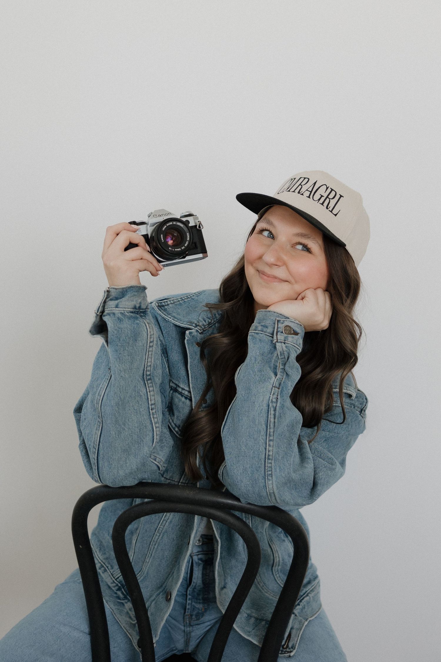 Smiling female photographer in denim jacket and cap holding vintage film camera while sitting on chair.