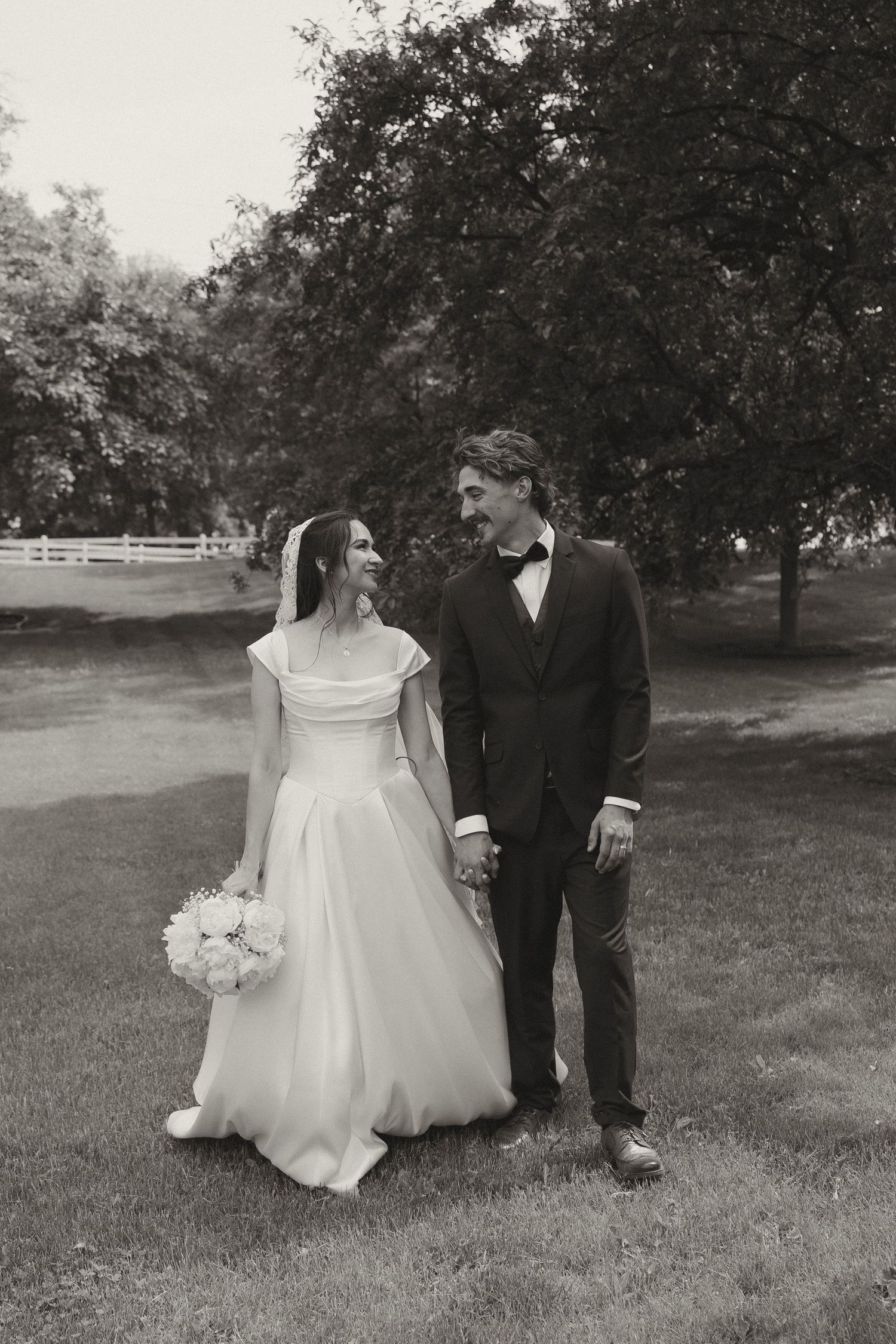 A couple walks through a scenic grassy field on their wedding day with trees in the background.