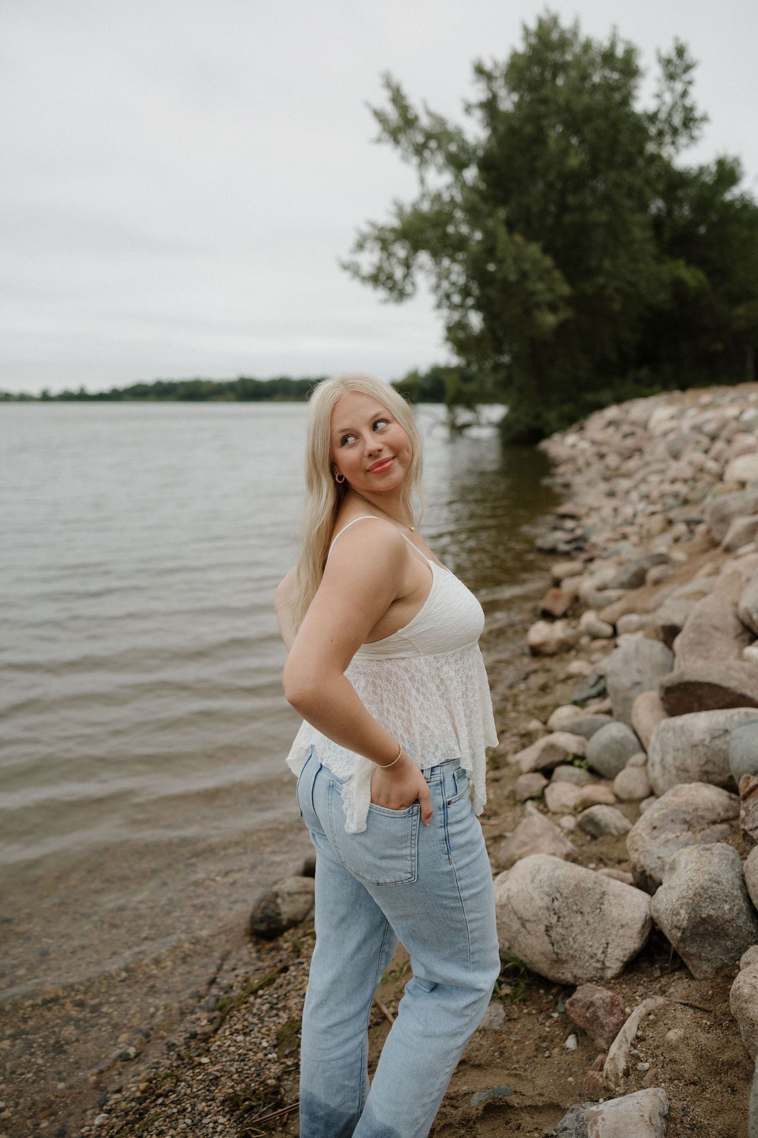 Blonde woman in white camisole and jeans standing on a rocky shoreline beside a calm lake with trees.