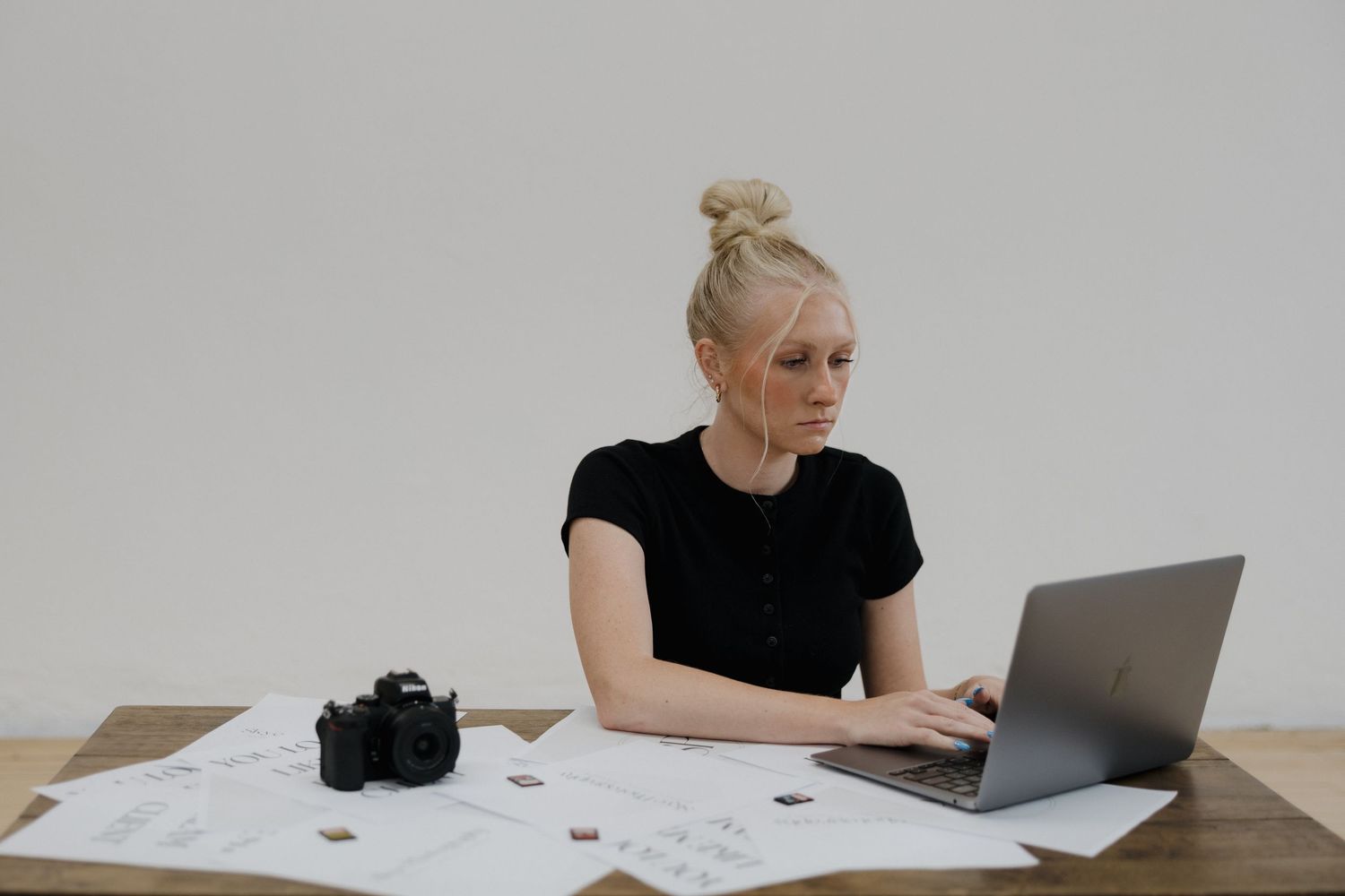 Blonde woman in black top working on laptop at wooden desk with camera and papers.