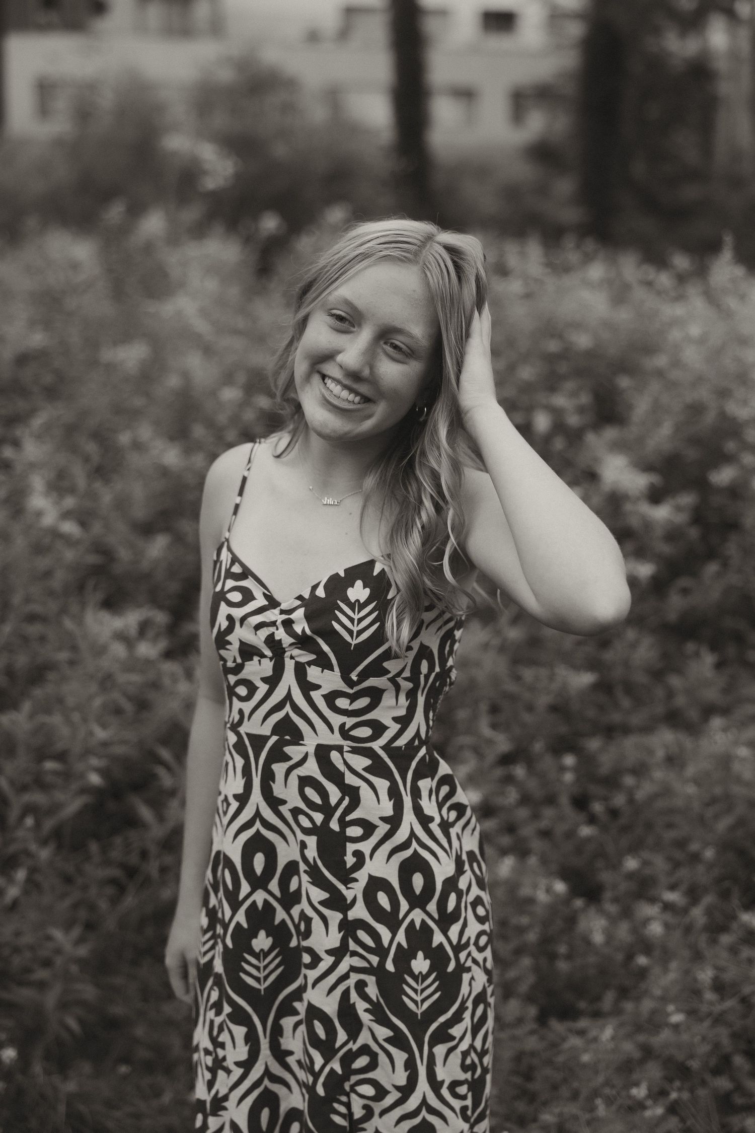 Black and white portrait of laughing blonde woman in patterned dress standing outdoors among lush green foliage.