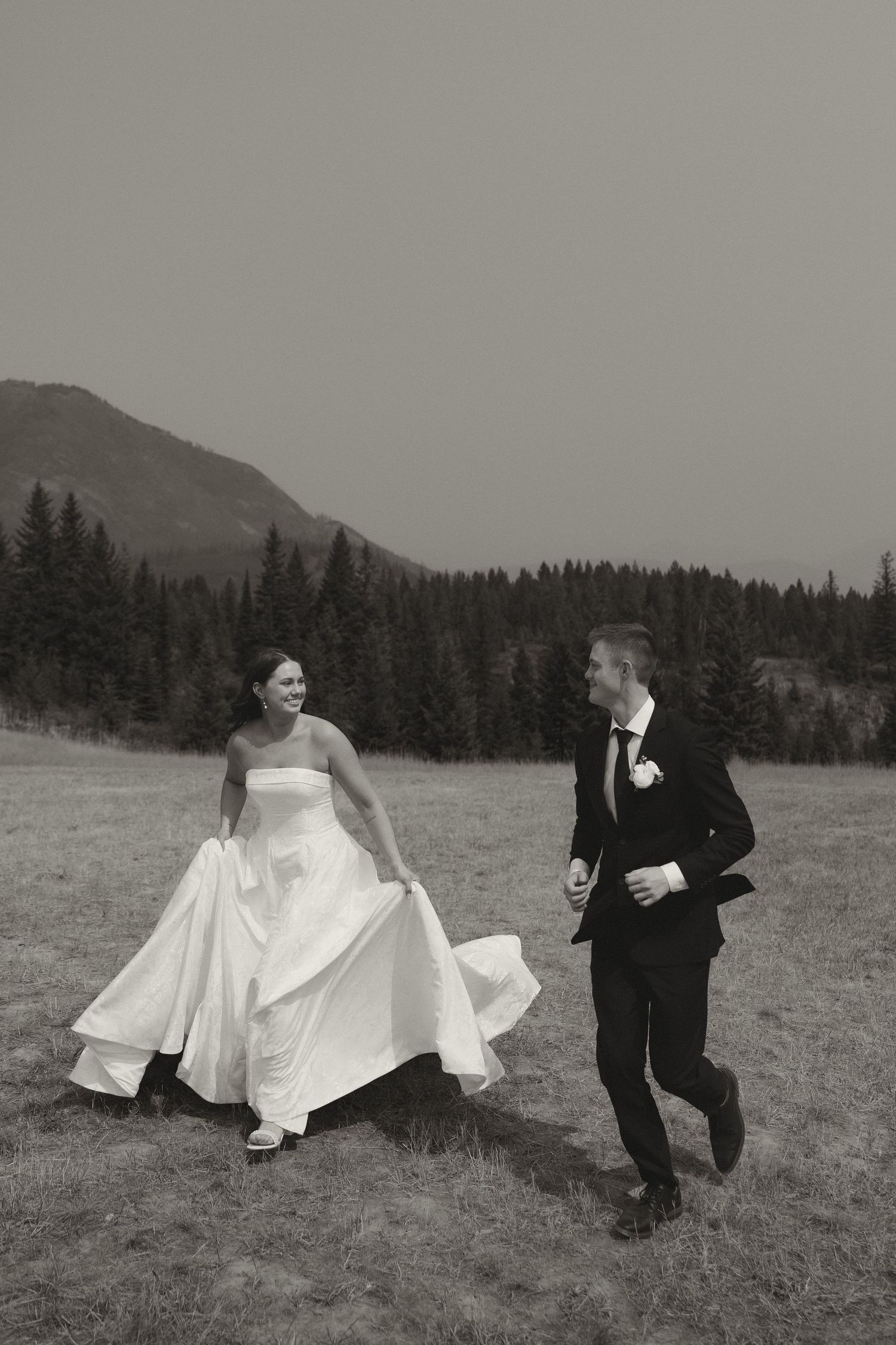 Black and white photo of a bride and groom running and laughing in a mountain meadow with pine trees.