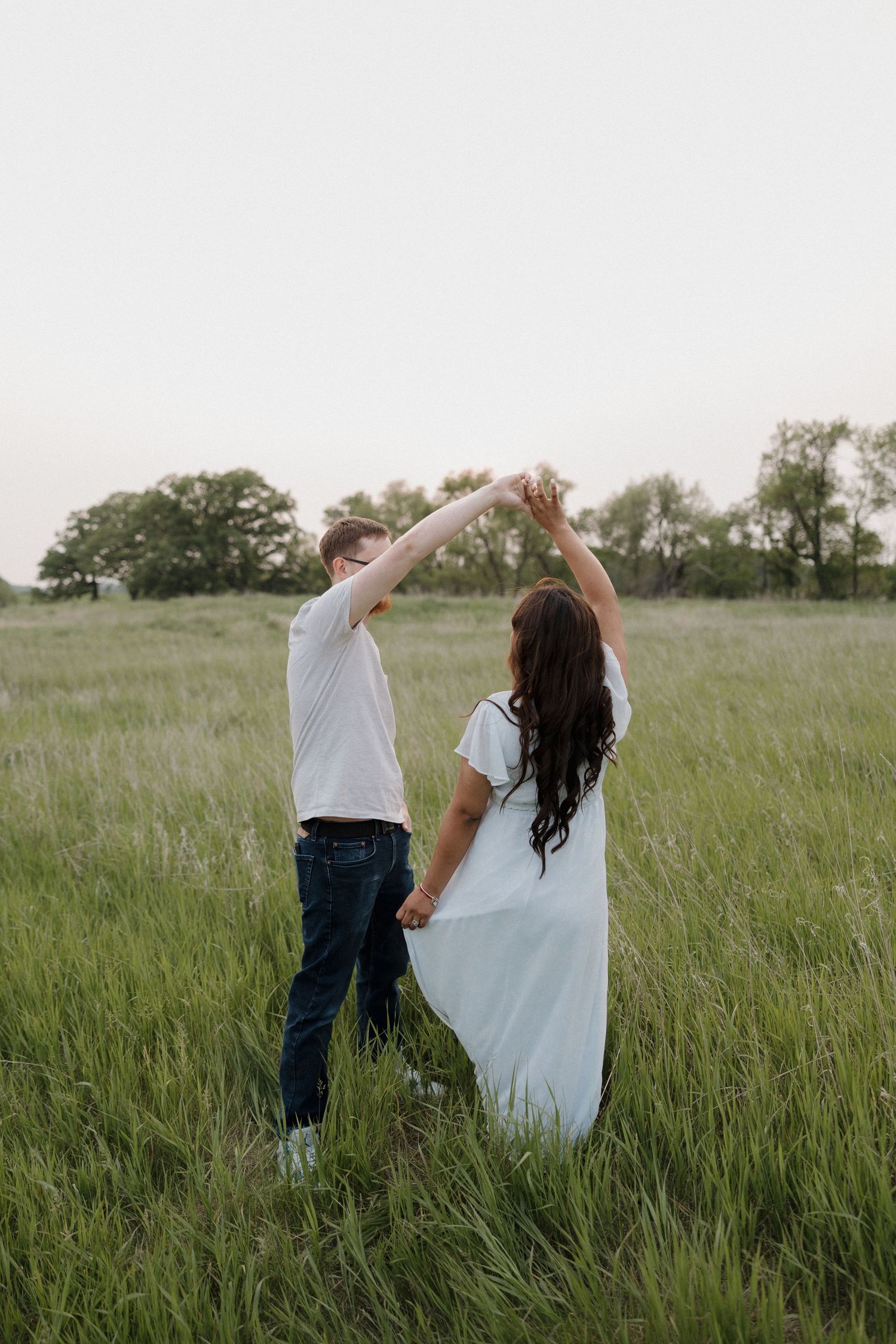 Couple dancing in a lush green field at dusk, holding hands raised, woman in white dress.