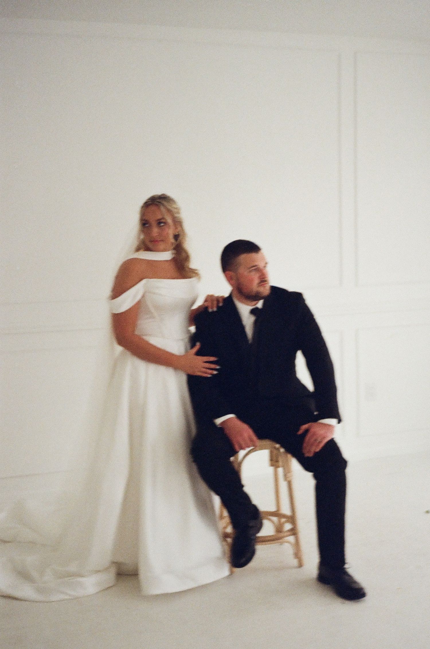 Bride in white off-shoulder gown stands beside groom in black tuxedo seated on gold chair in bright white studio.