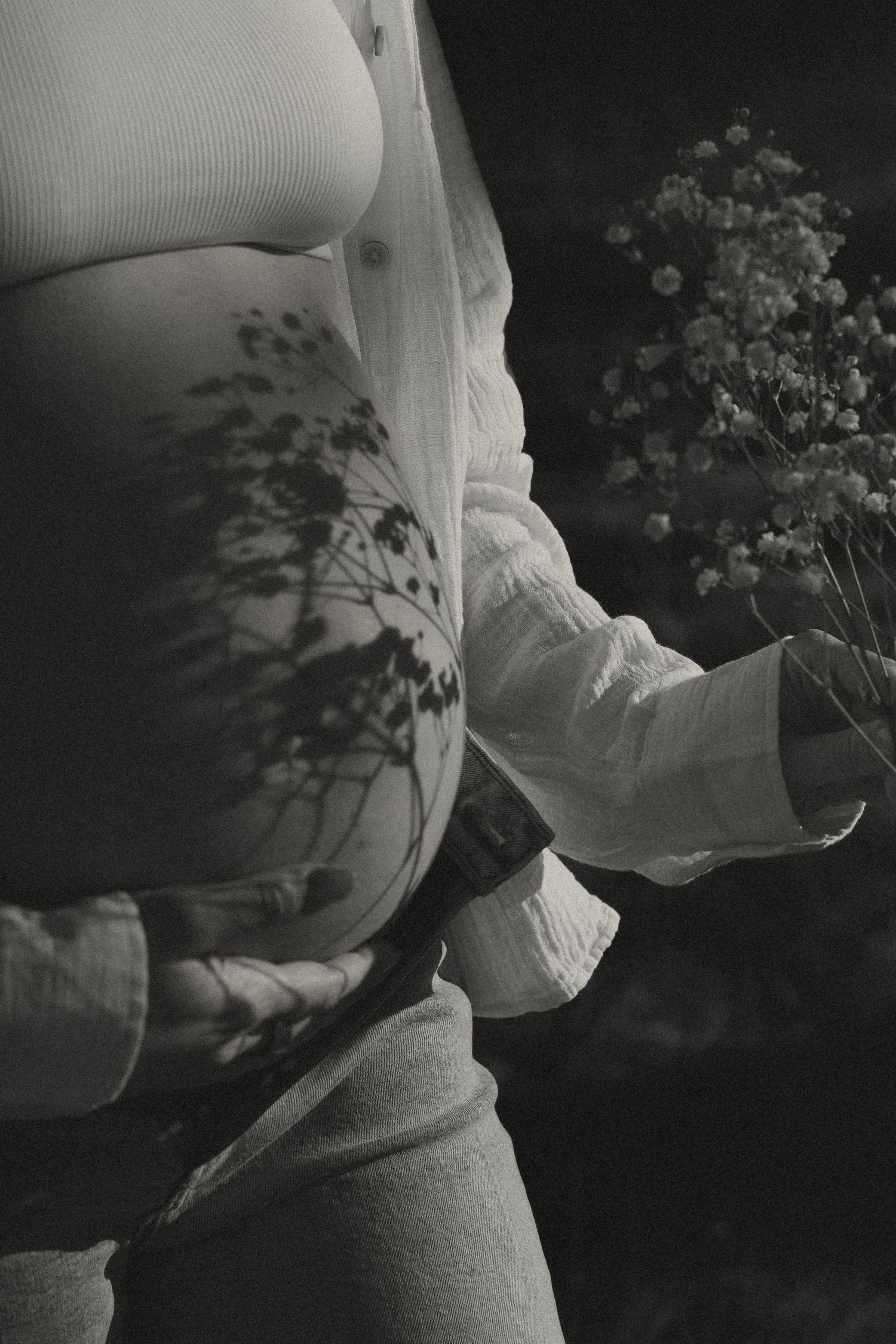 Black and white close-up of pregnant belly with floral shadow, woman holding wildflower bouquet outdoors.