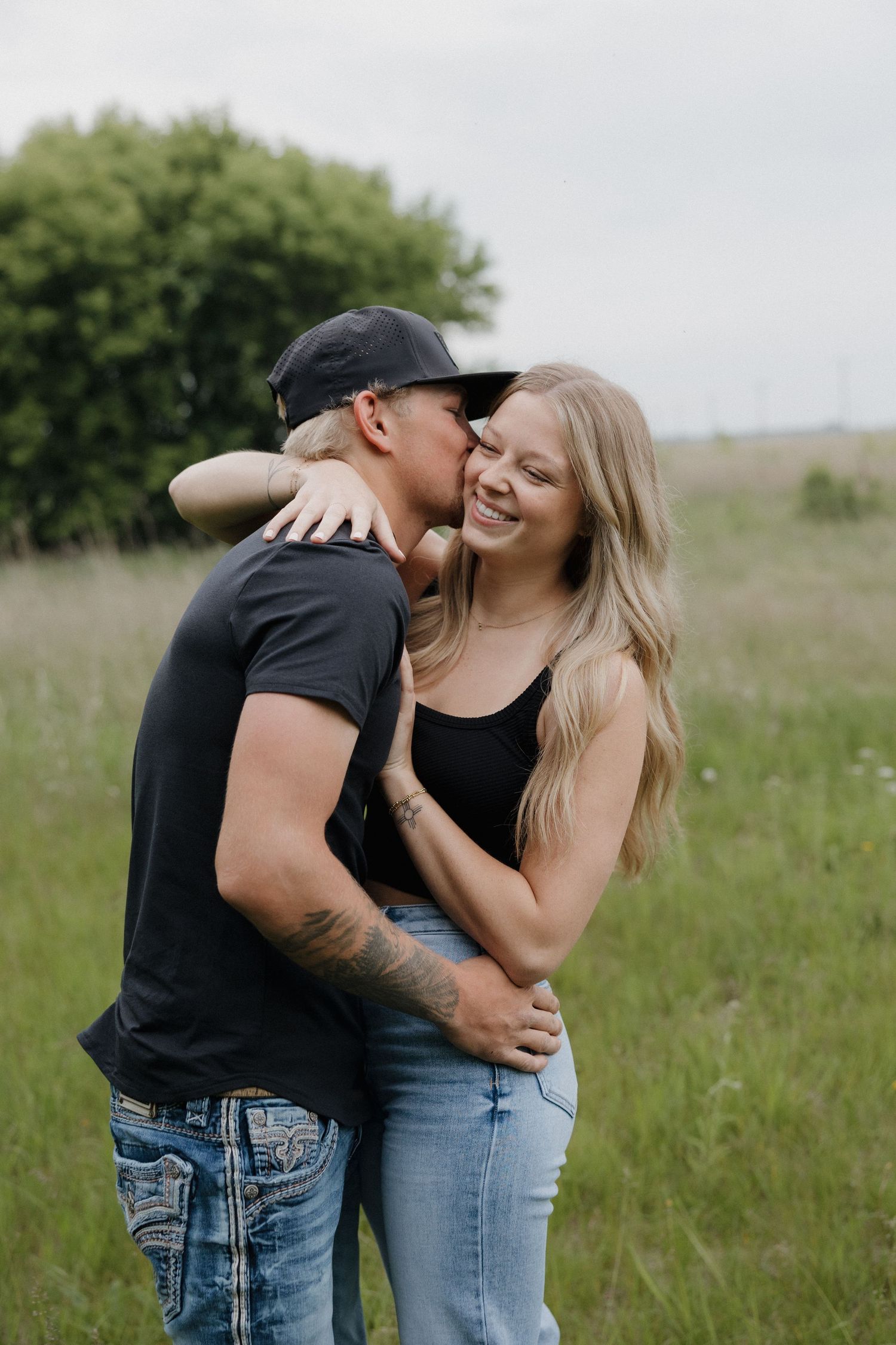 Tattooed man in black cap kissing smiling blonde woman's cheek in lush green field.