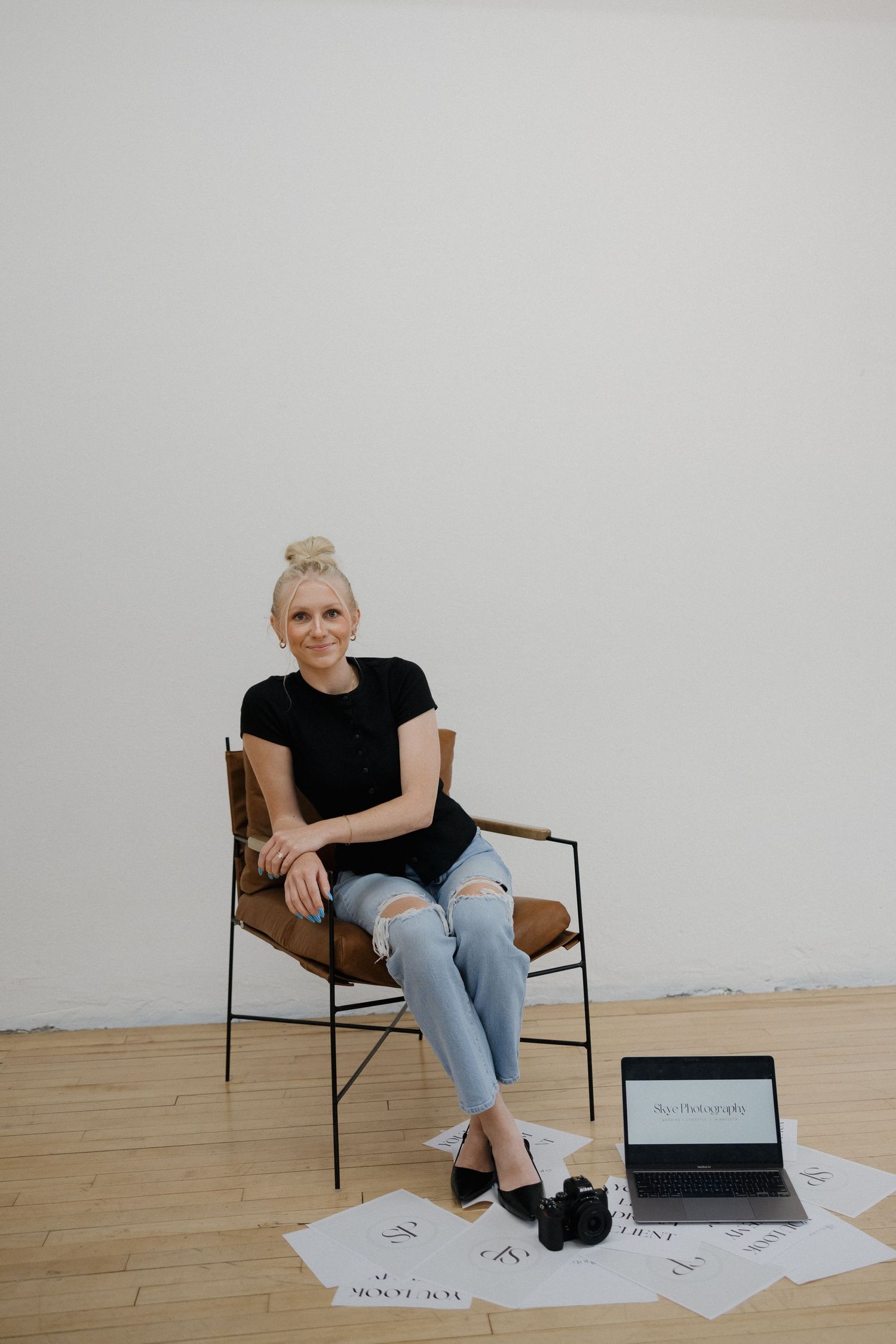 Blonde woman in black tee and ripped jeans sitting in leather chair with laptop and camera.