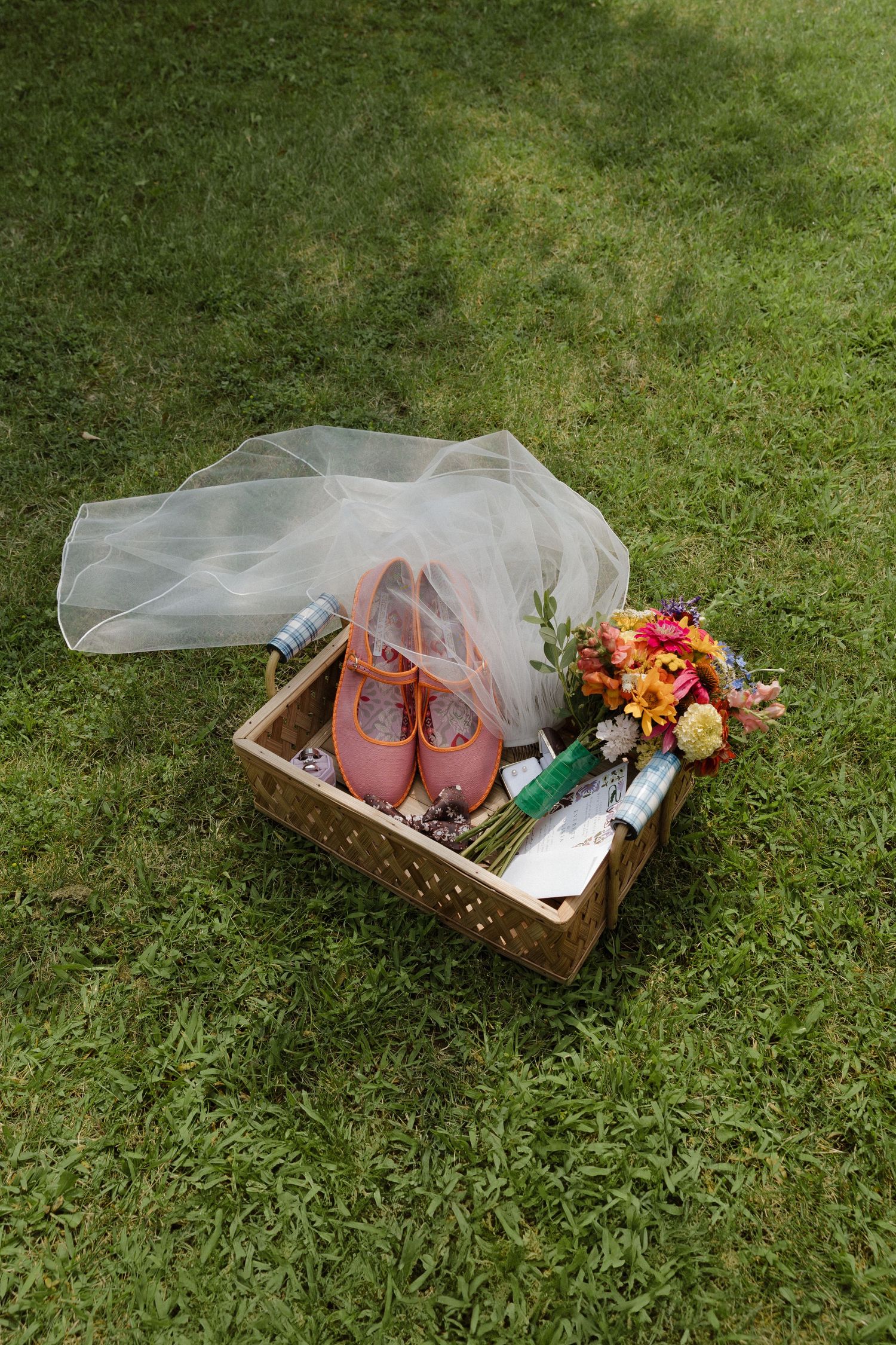 A rustic wooden box containing wedding accessories with pink shoes, flowers, and a veil on grass.
