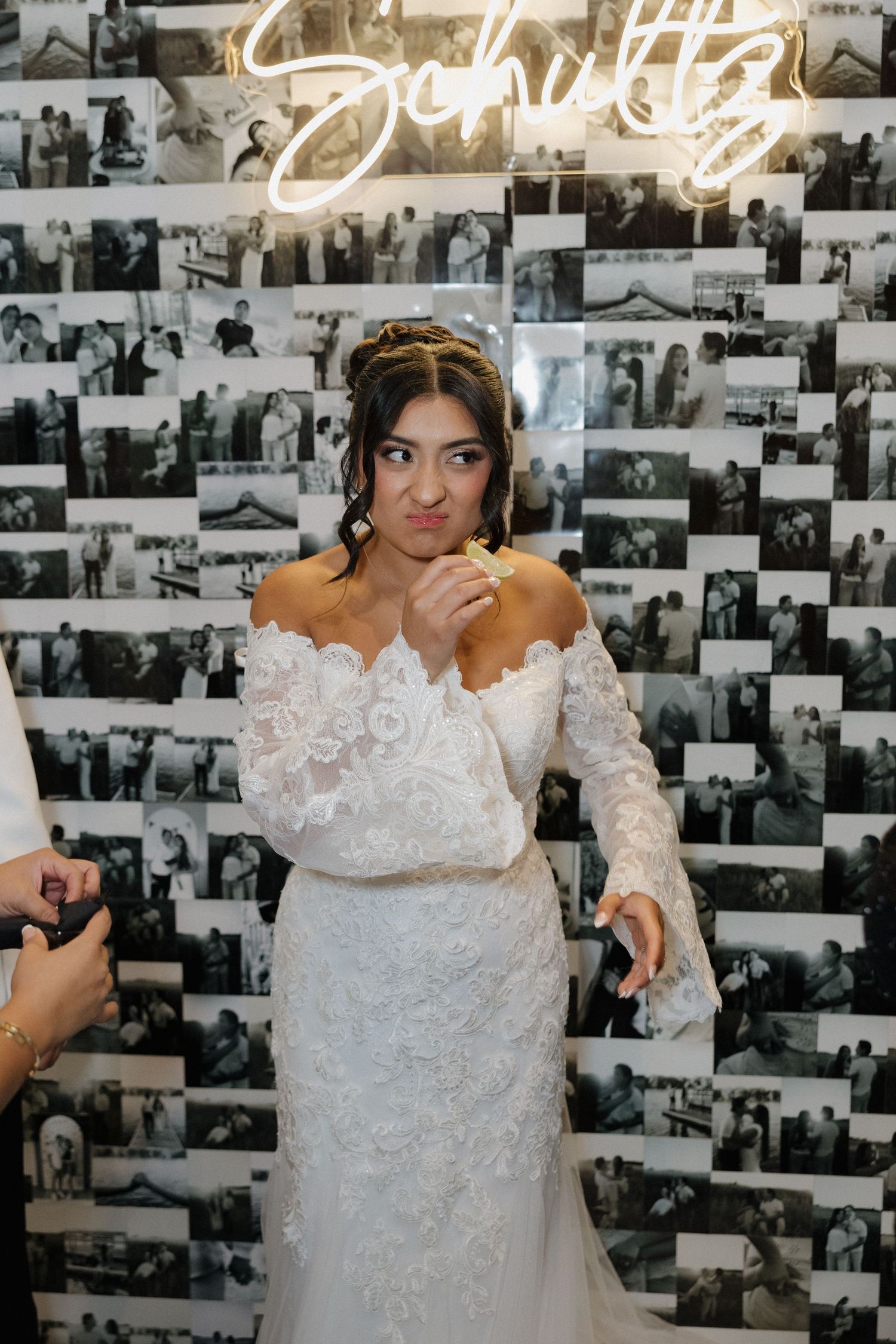 A bride in a lace wedding dress poses against a wall decorated with black and white photographs.