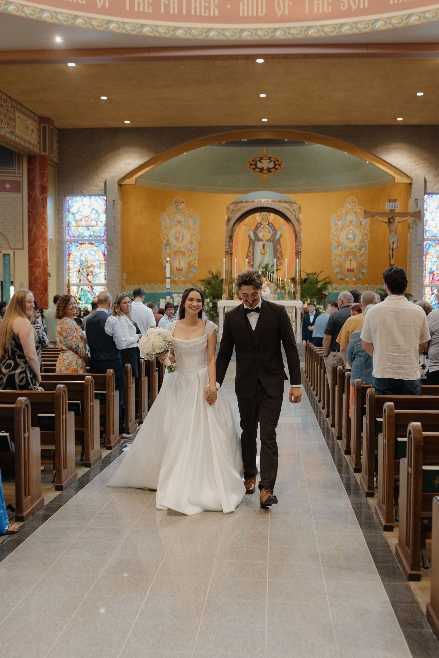 Wedding ceremony procession inside a traditional church with stained glass windows and wooden pews.