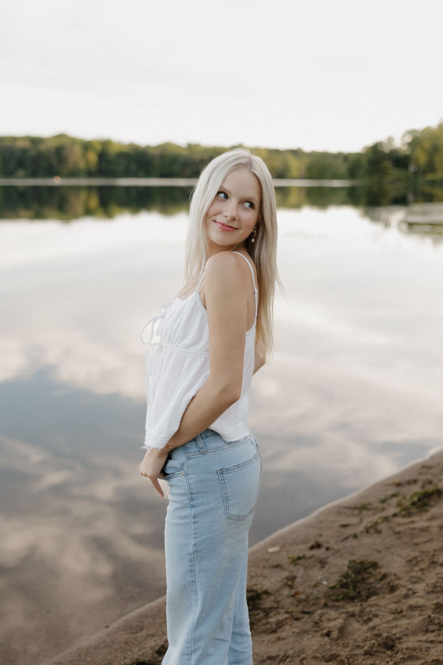 Blonde woman in white top and jeans posing by a calm reflective lake at golden hour.