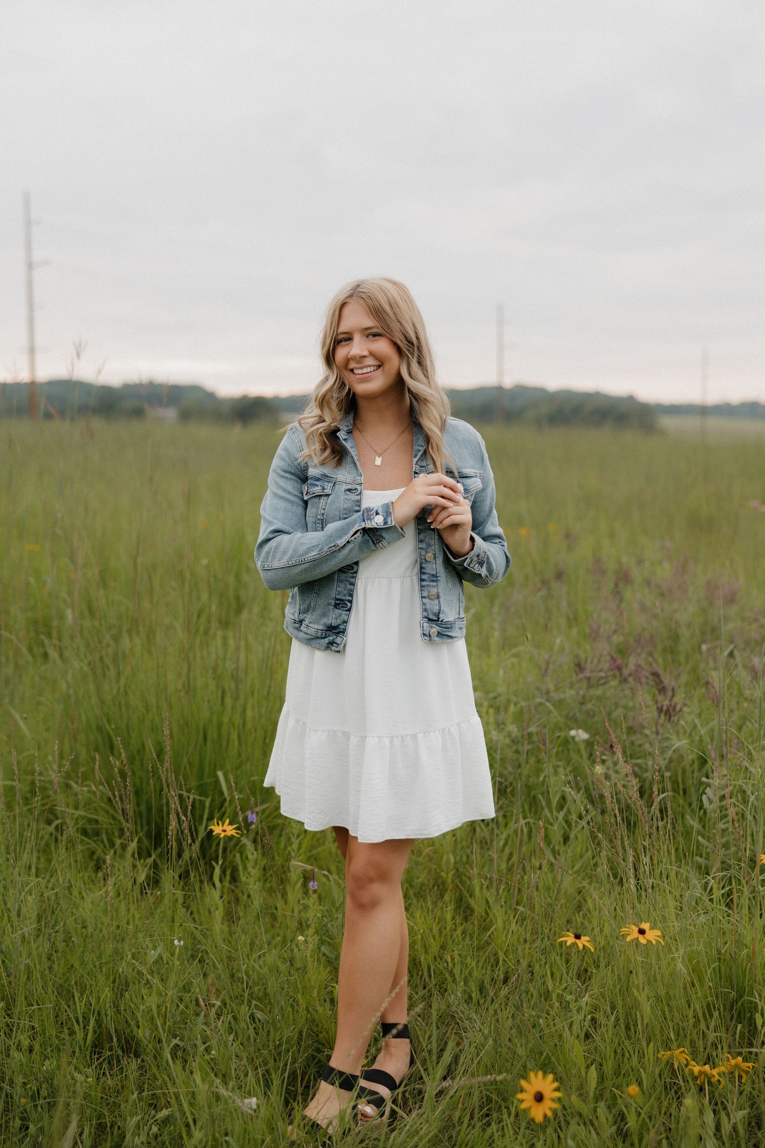 Smiling blonde woman in white dress and denim jacket standing in a wildflower meadow.