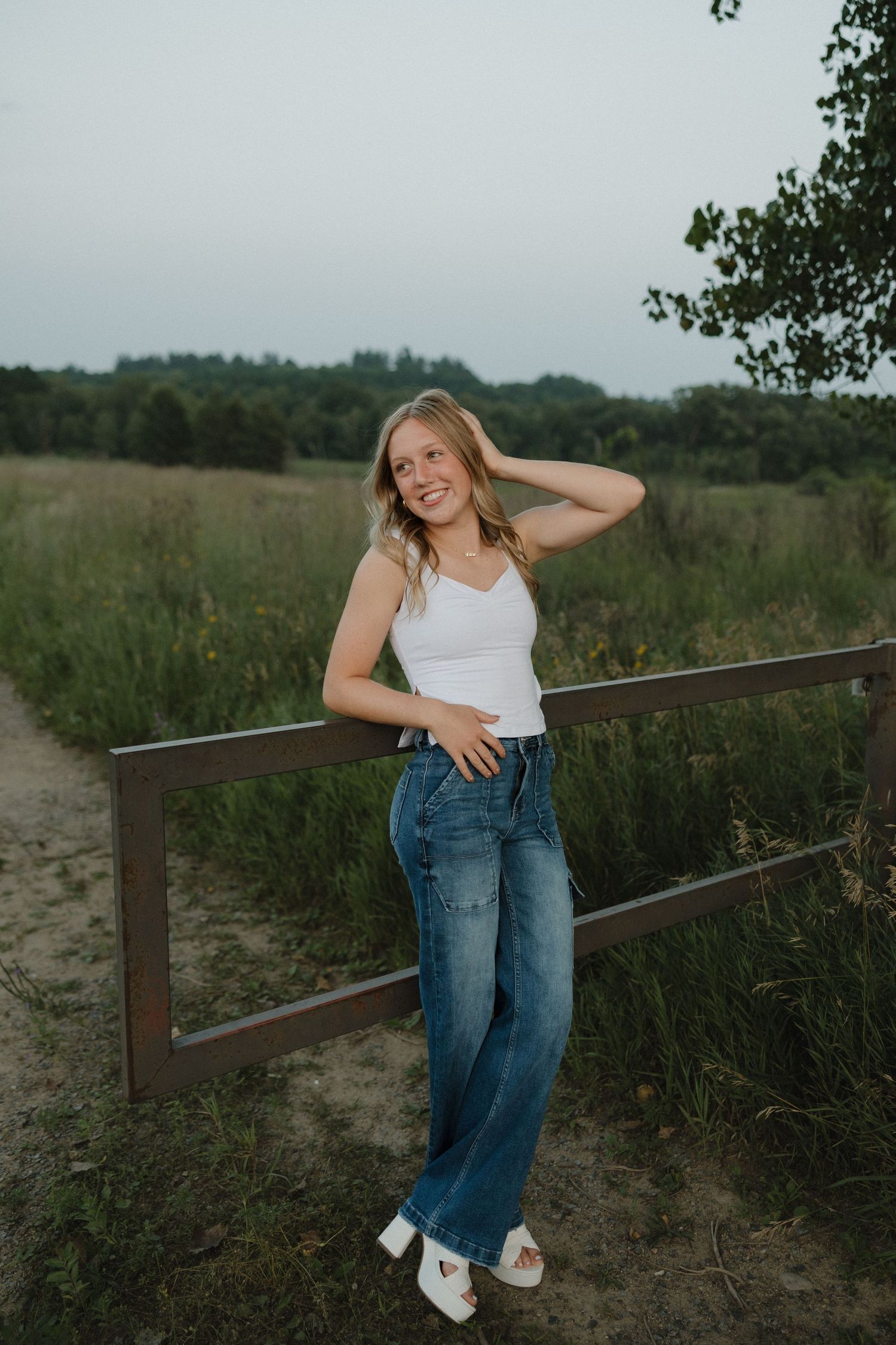 Smiling blonde woman in white tank top and wide-leg jeans leaning on wooden fence in green meadow.