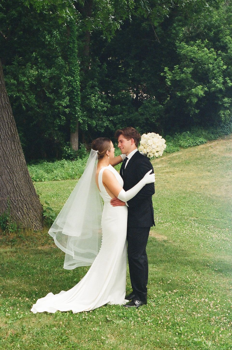 Bride and groom share a romantic moment outdoors, with bride in white gown and veil holding bouquet.