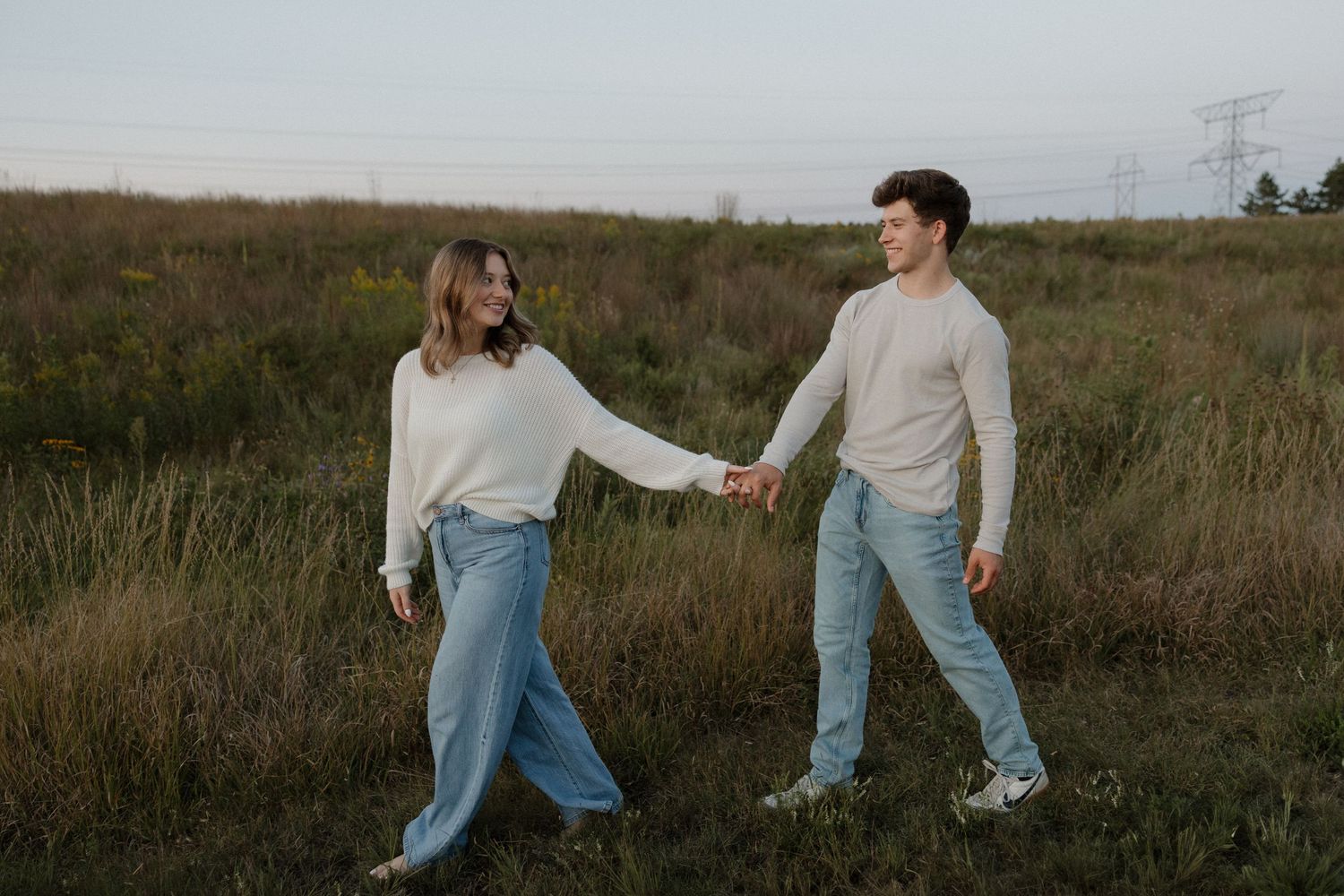 Young couple holding hands and walking through an open field at dusk, both wearing jeans and light sweaters.