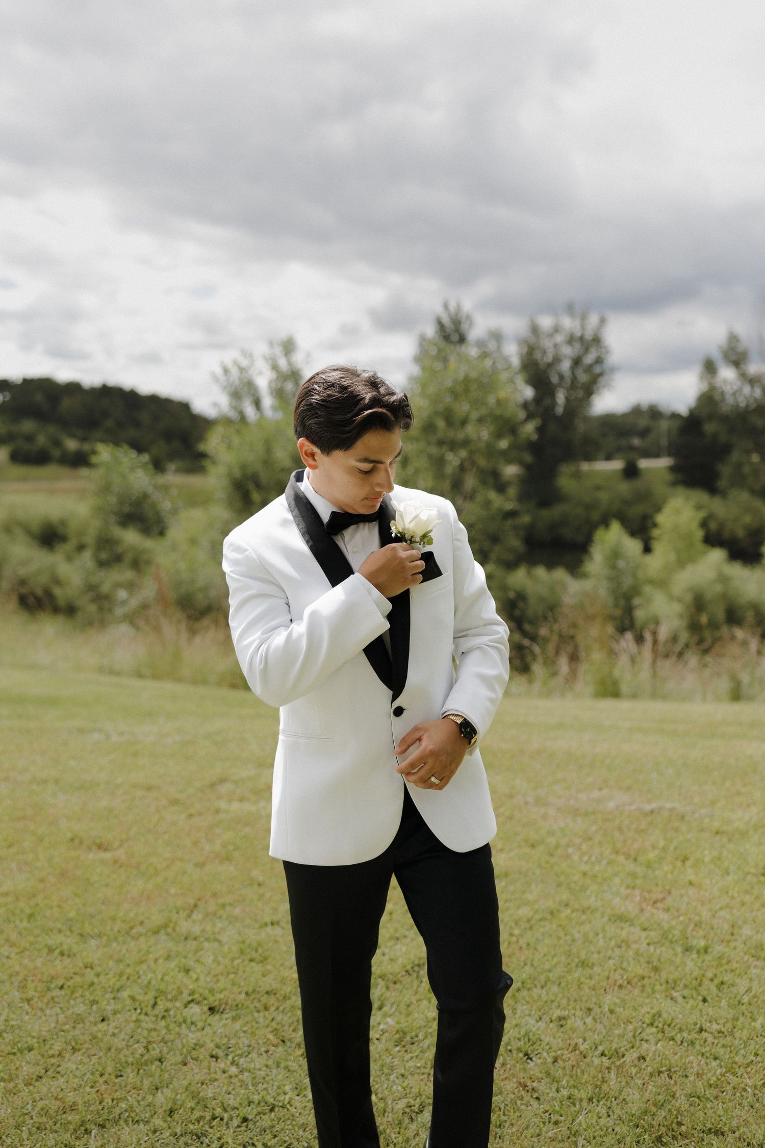A person in a white tuxedo jacket and black bow tie wipes away tears in an outdoor field setting.