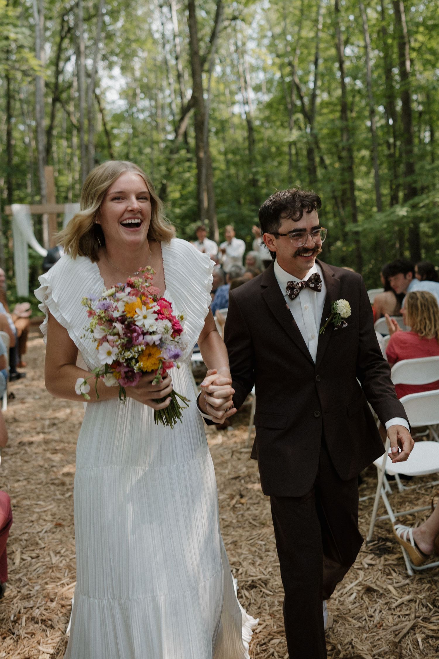 Joyful wedding ceremony exit in wooded setting with colorful bouquet.