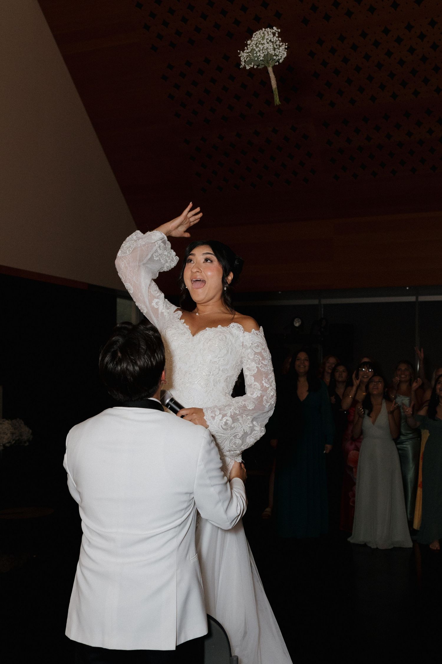 A wedding bouquet toss moment captured during a reception in a dimly lit venue.