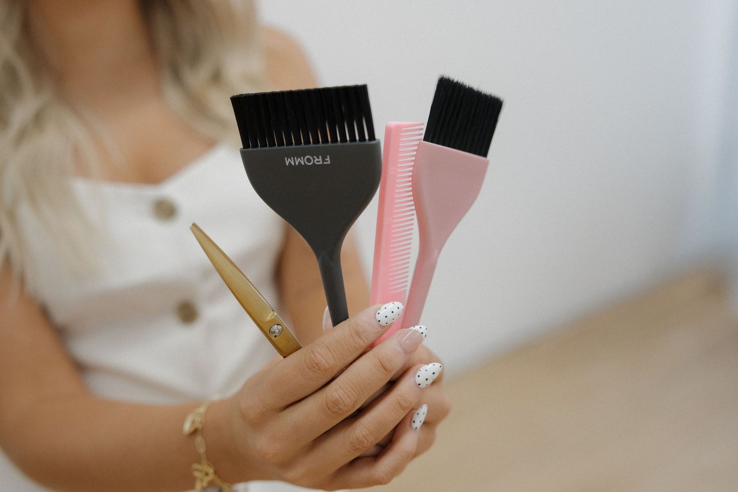 Blonde woman holding three hair coloring brushes in black and pink in a bright room.