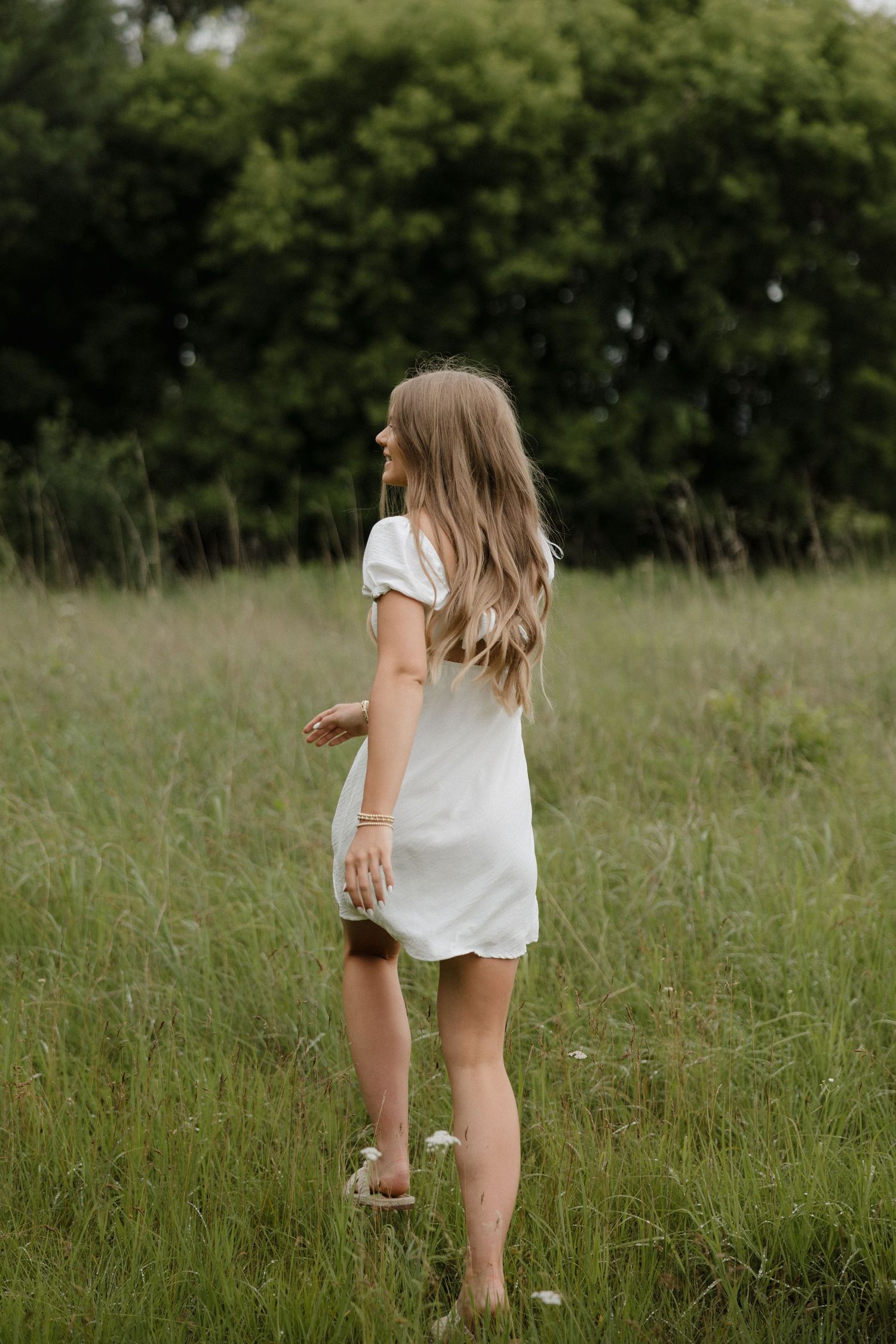 Young woman in white dress standing barefoot in a lush green field facing away.