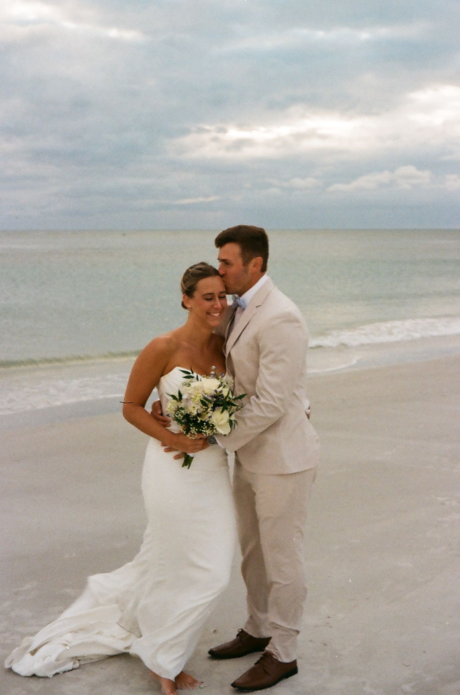 Bride and groom embrace on a sandy beach, she holds a white bouquet, he wears a tan suit, ocean waves in the background.