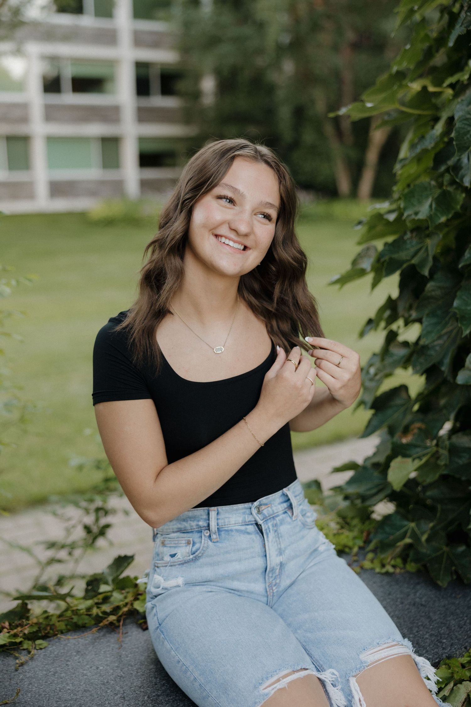Smiling teenage girl in black top and denim shorts poses beside green ivy in outdoor setting.