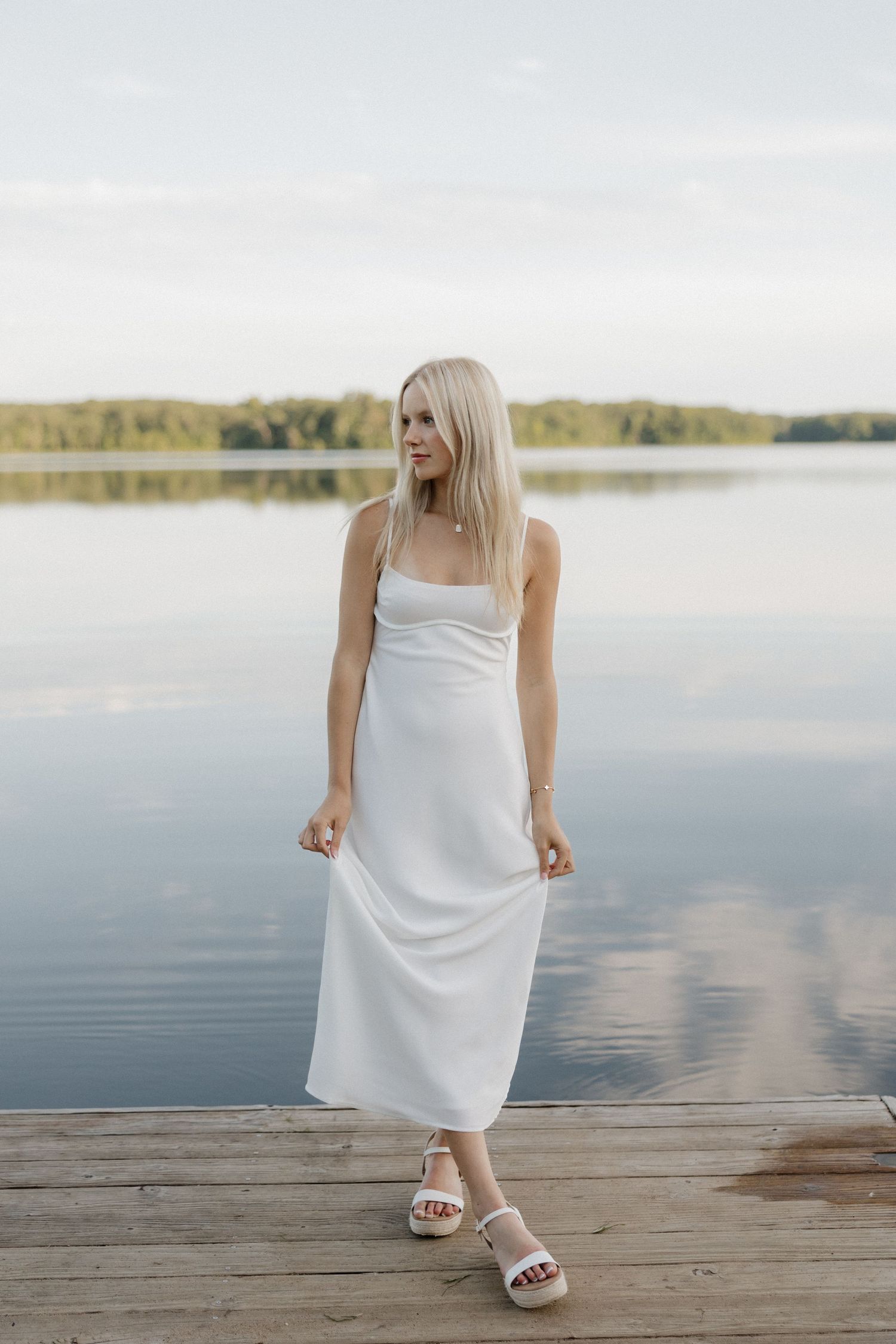 Blonde woman in white slip midi dress standing on a wooden dock overlooking a calm lake.